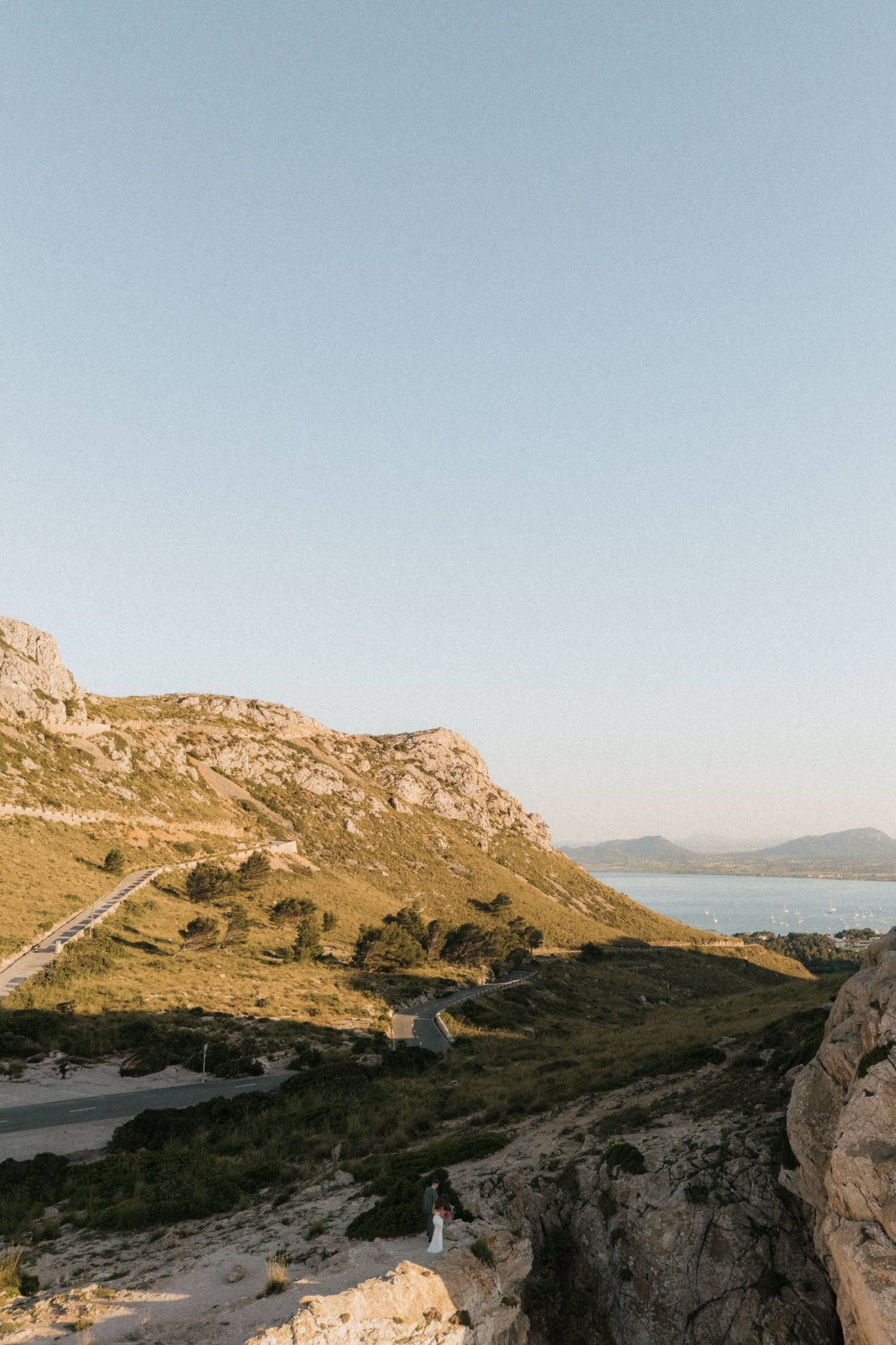 Scenic coastal landscape with rocky mountains, green hills, and calm blue waters under a clear sky at golden hour.