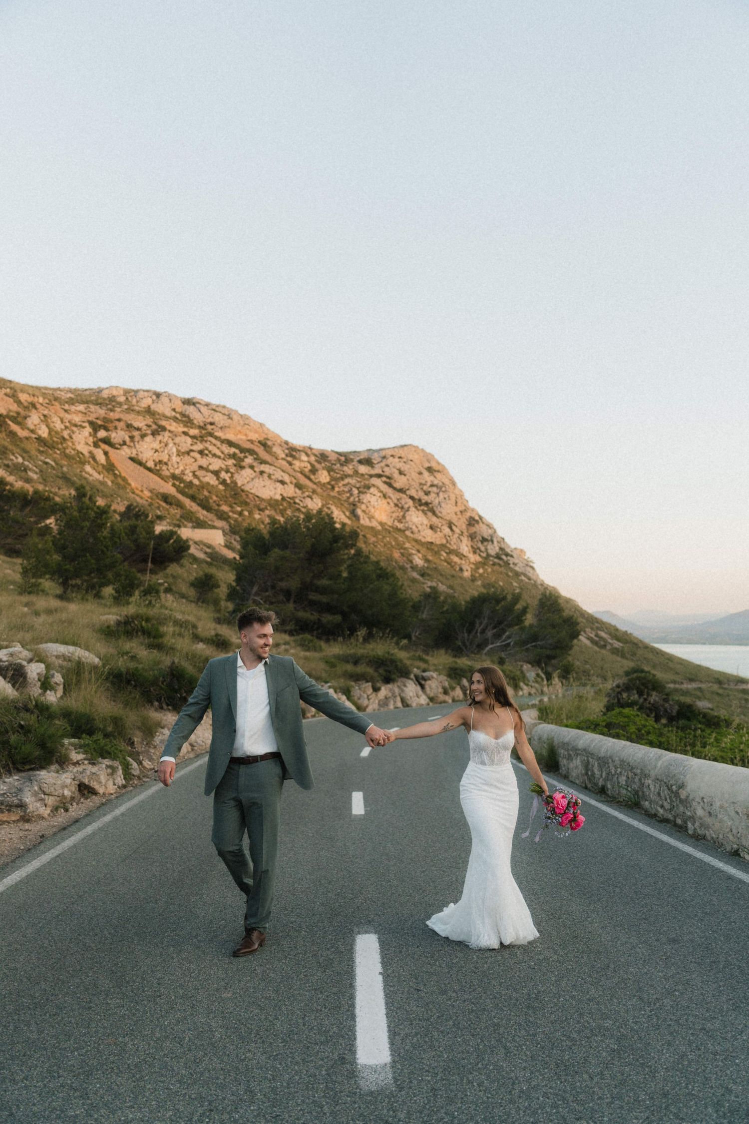 Bride and groom walk hand-in-hand down a scenic coastal road with mountains at sunset.