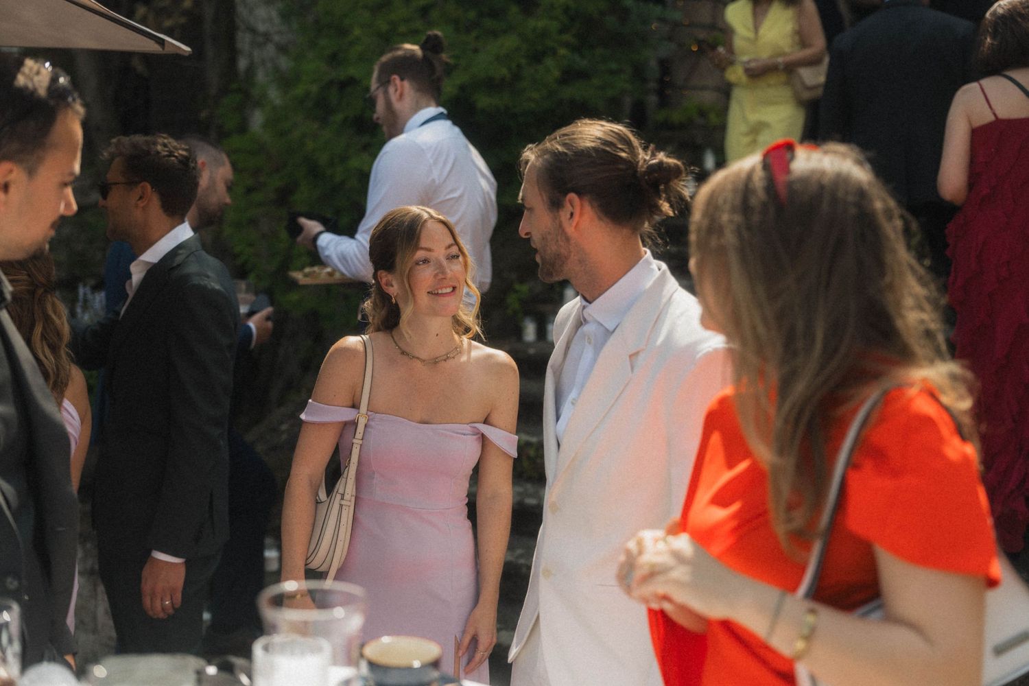 Wedding guests mingling outdoors, woman in pink dress conversing with man in white suit.