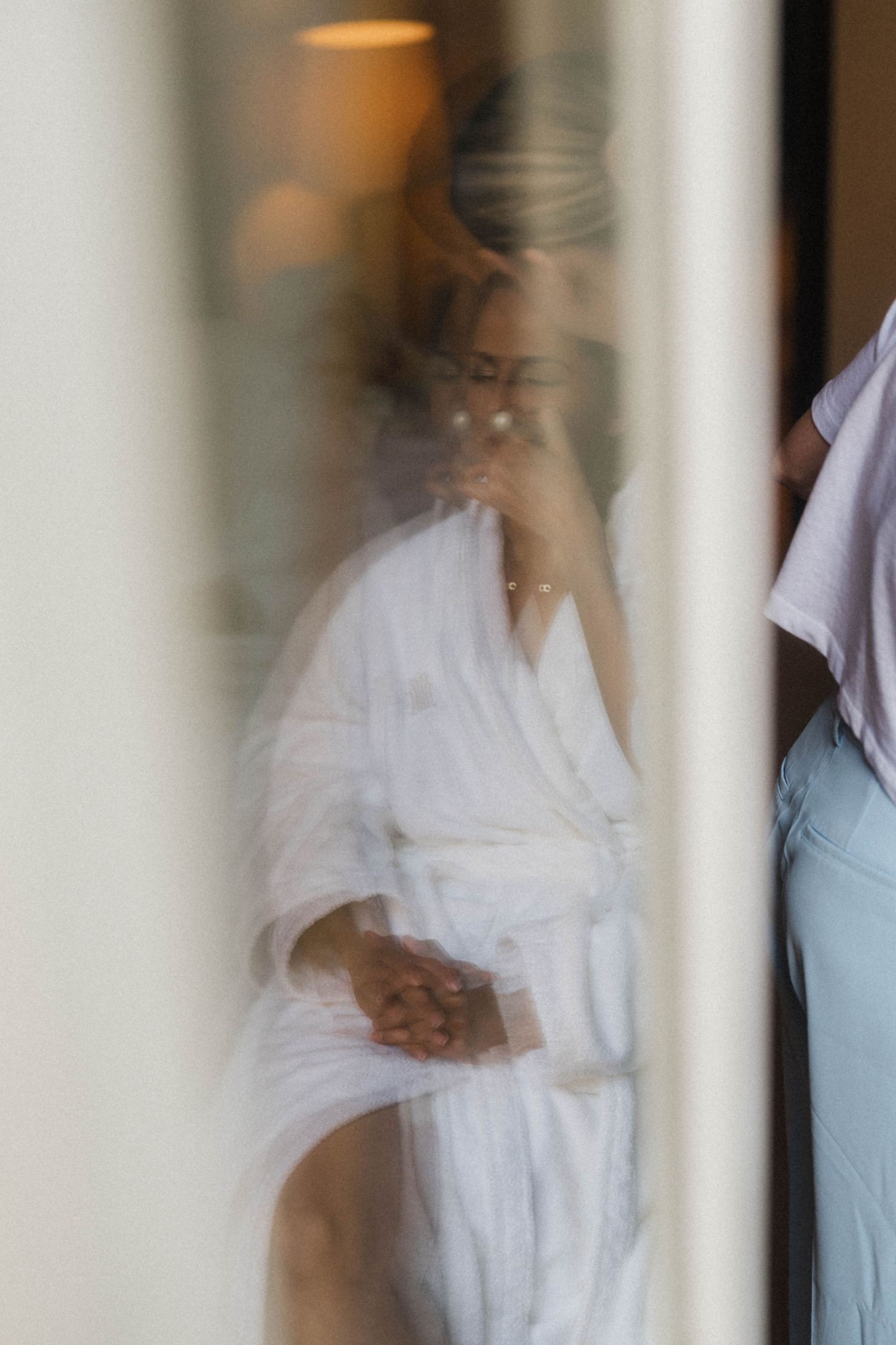 Bride in white robe preparing for wedding day, seen through glass reflection.