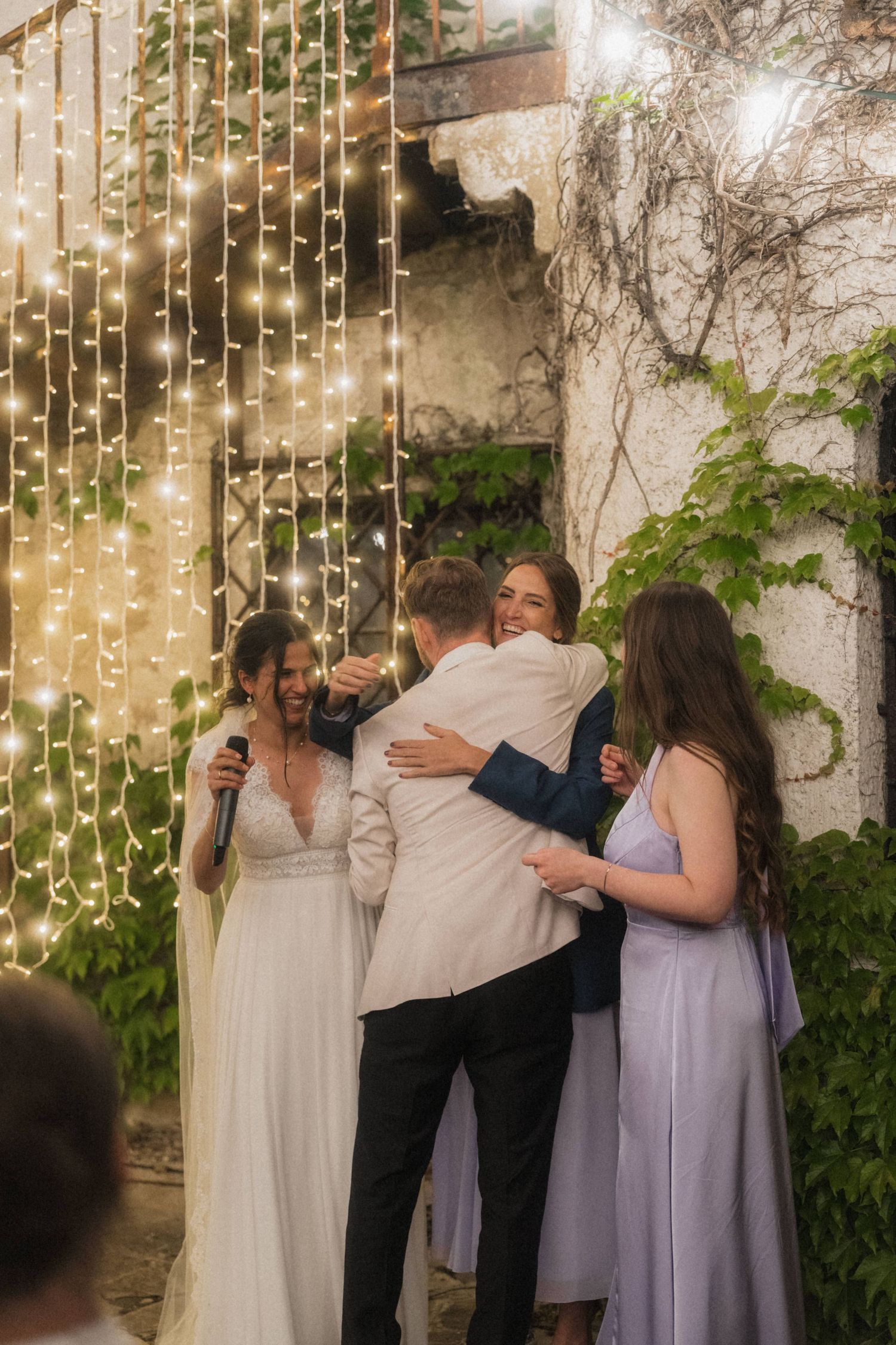 Groom embracing guest near bride at evening wedding reception with fairy light curtain backdrop.