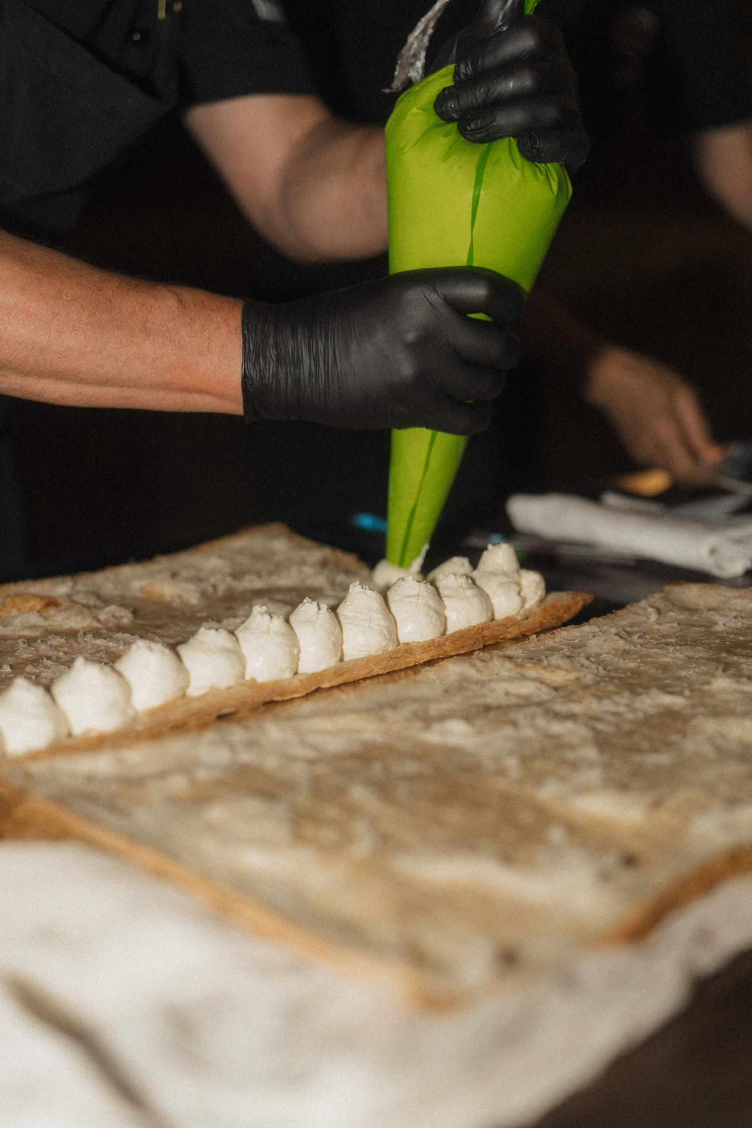Chef in black gloves piping cream filling onto pastry sheet in professional kitchen.