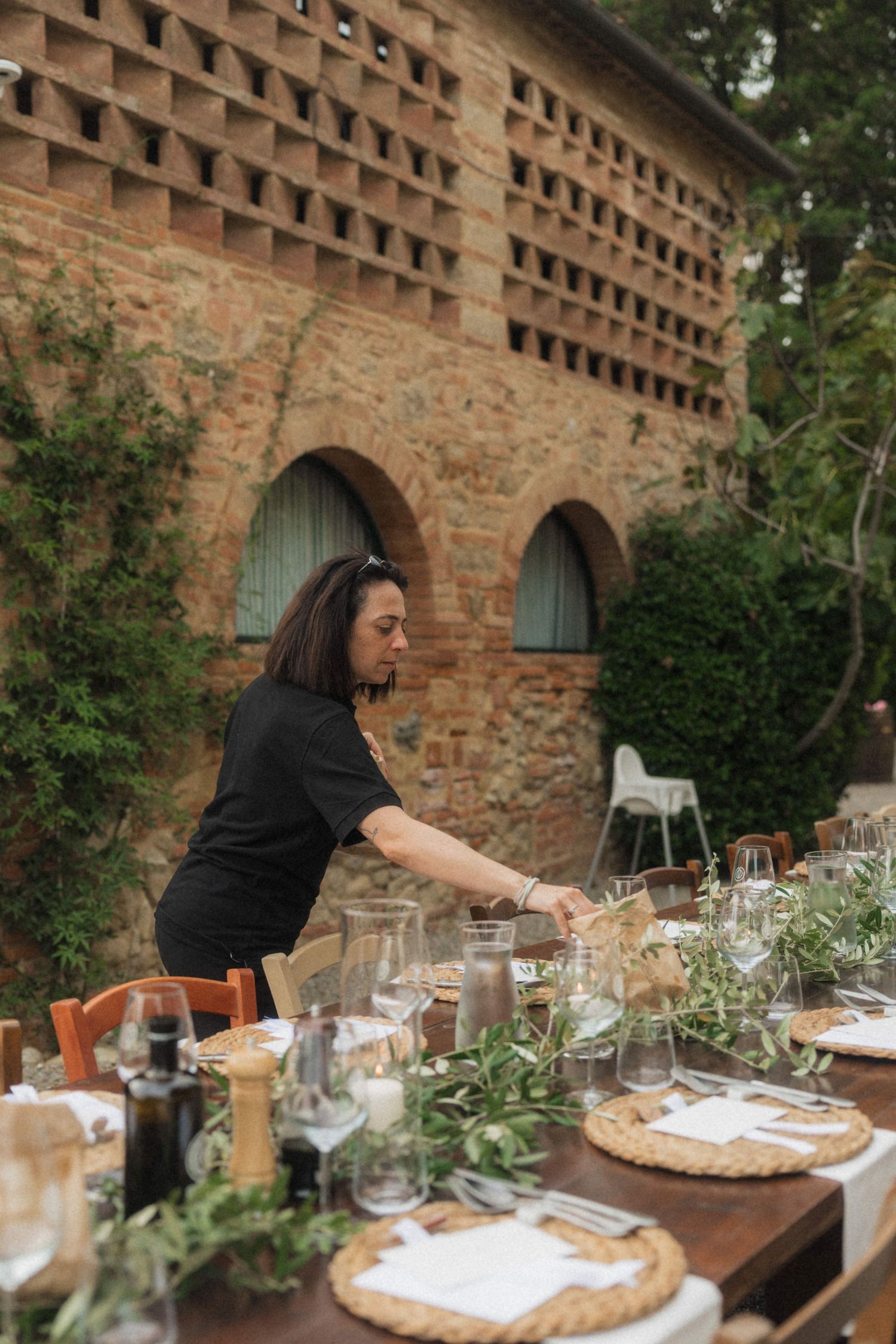 Woman in black dress setting an elegant outdoor dining table with glassware and greenery at a rustic stone venue.