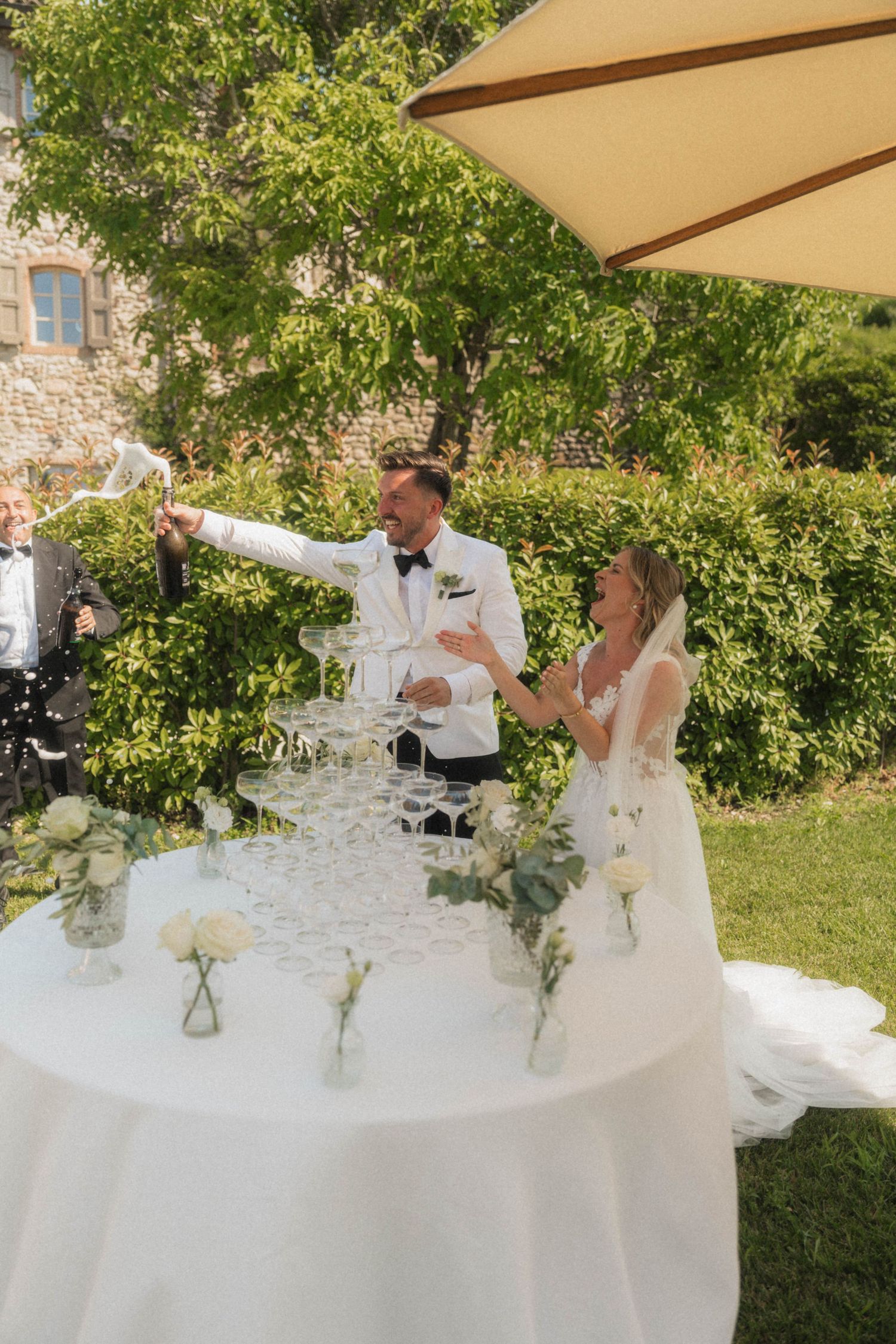 Bride and groom pouring champagne tower at outdoor wedding reception with white floral decorations.