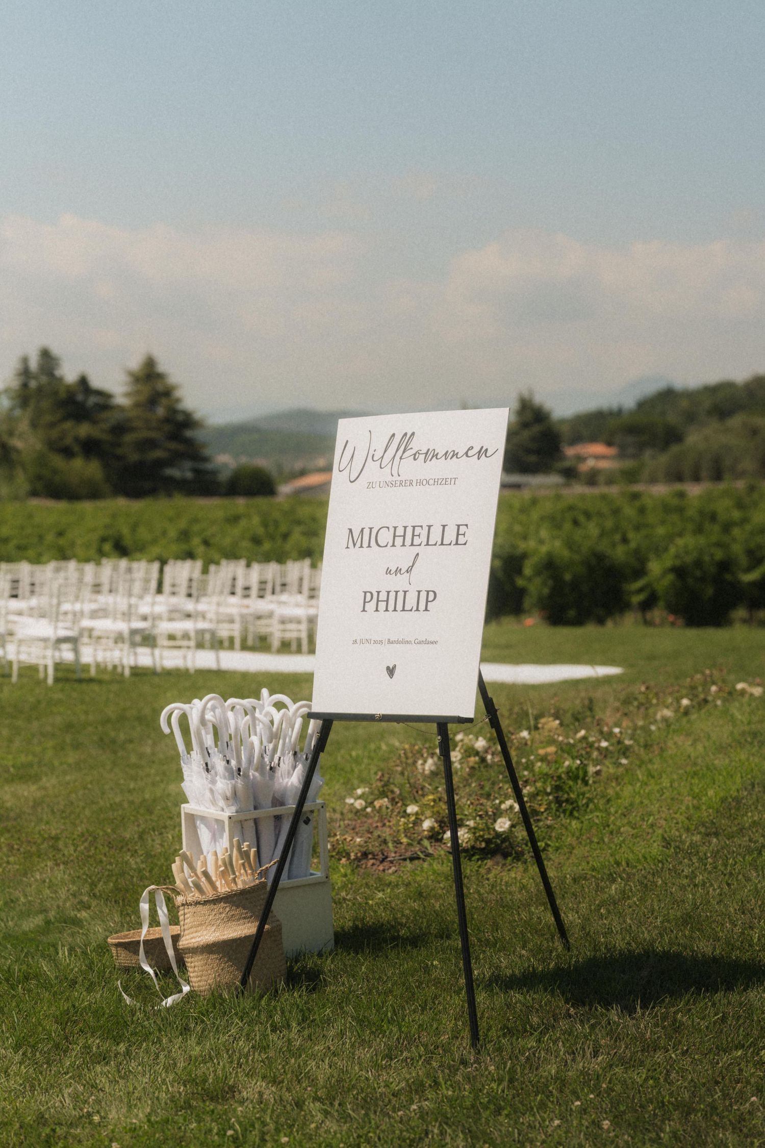 Outdoor wedding welcome sign for Michelle and Philip on an easel with white umbrellas on a sunny vineyard lawn.