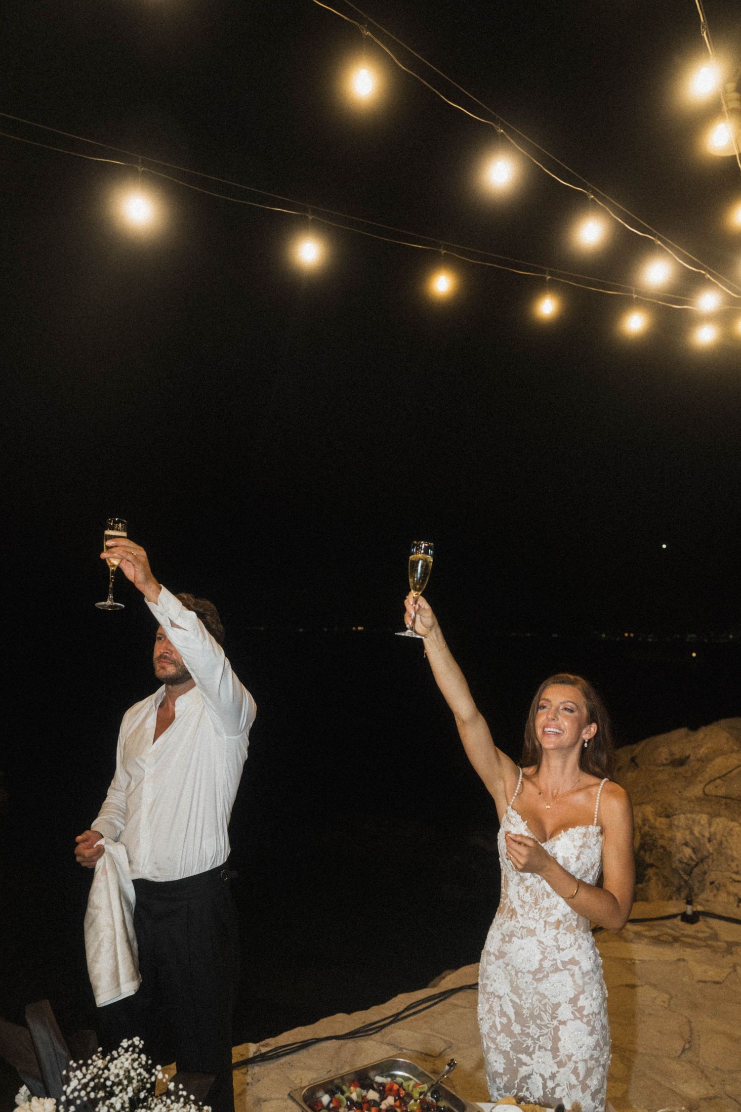 Bride and groom raising champagne glasses under string lights at romantic outdoor nighttime wedding celebration.