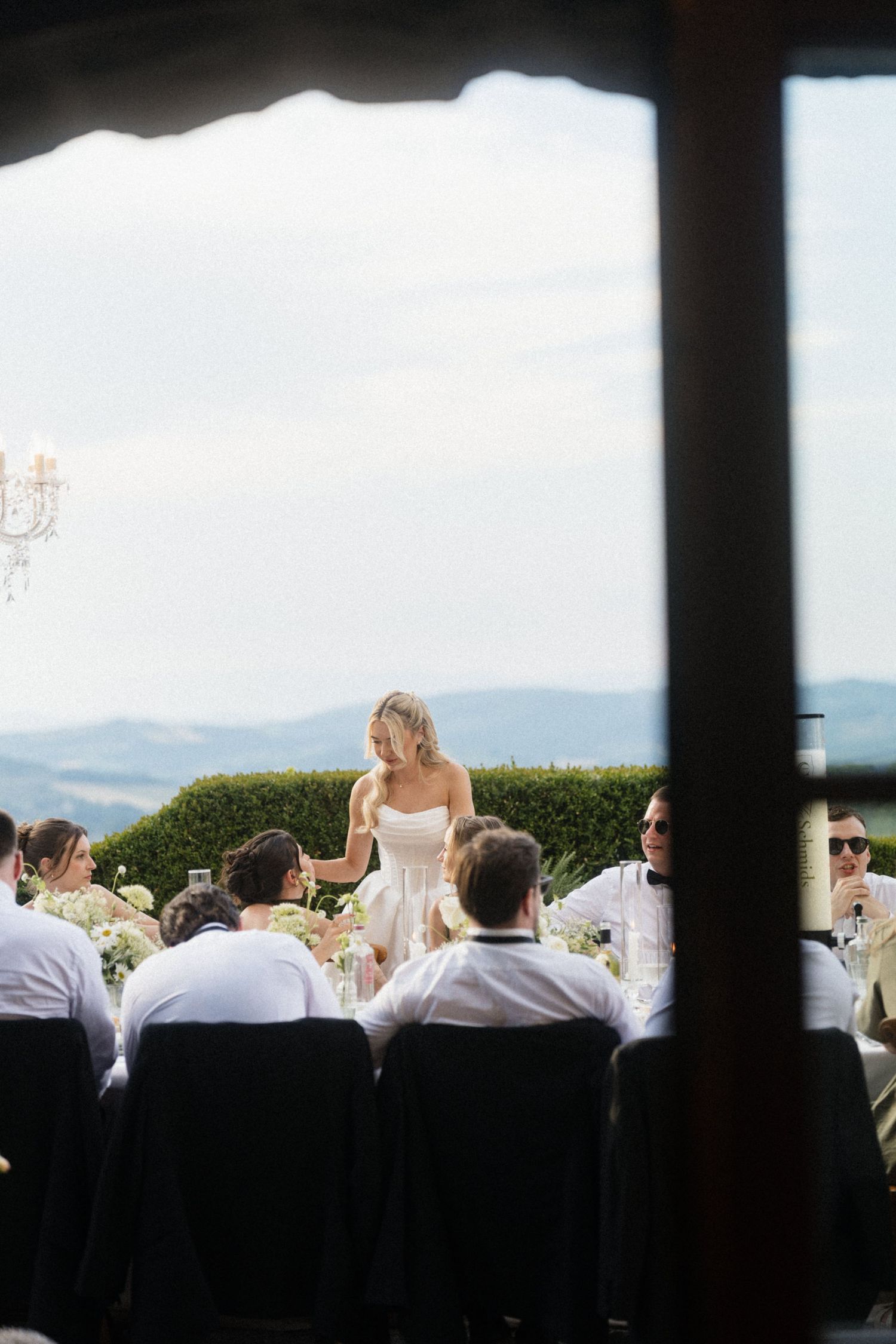 Bride in strapless white gown stands at outdoor reception table with panoramic Tuscan countryside backdrop.