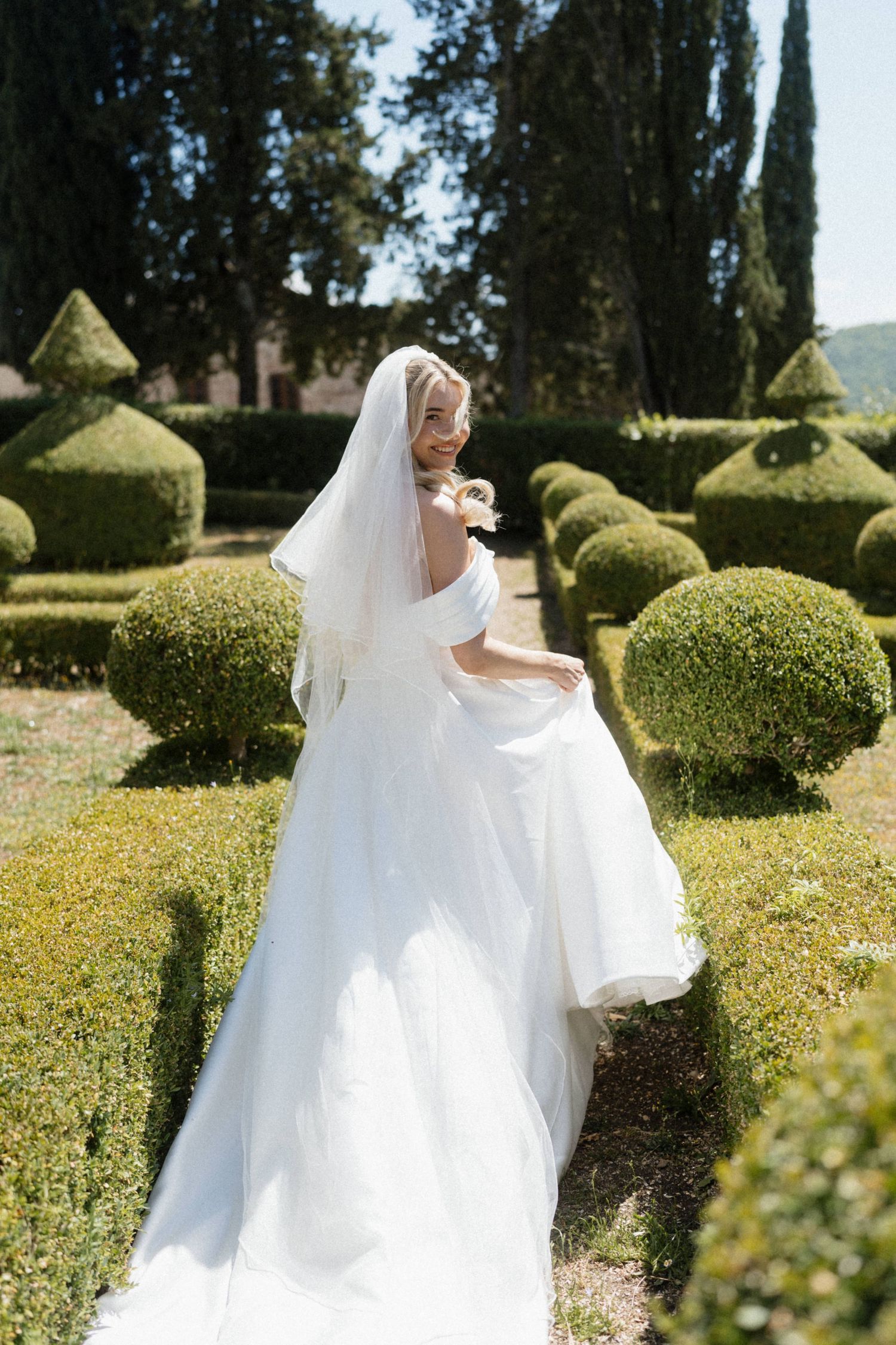 Bride in off-shoulder white gown and veil twirls among manicured topiary hedges in formal Italian garden.