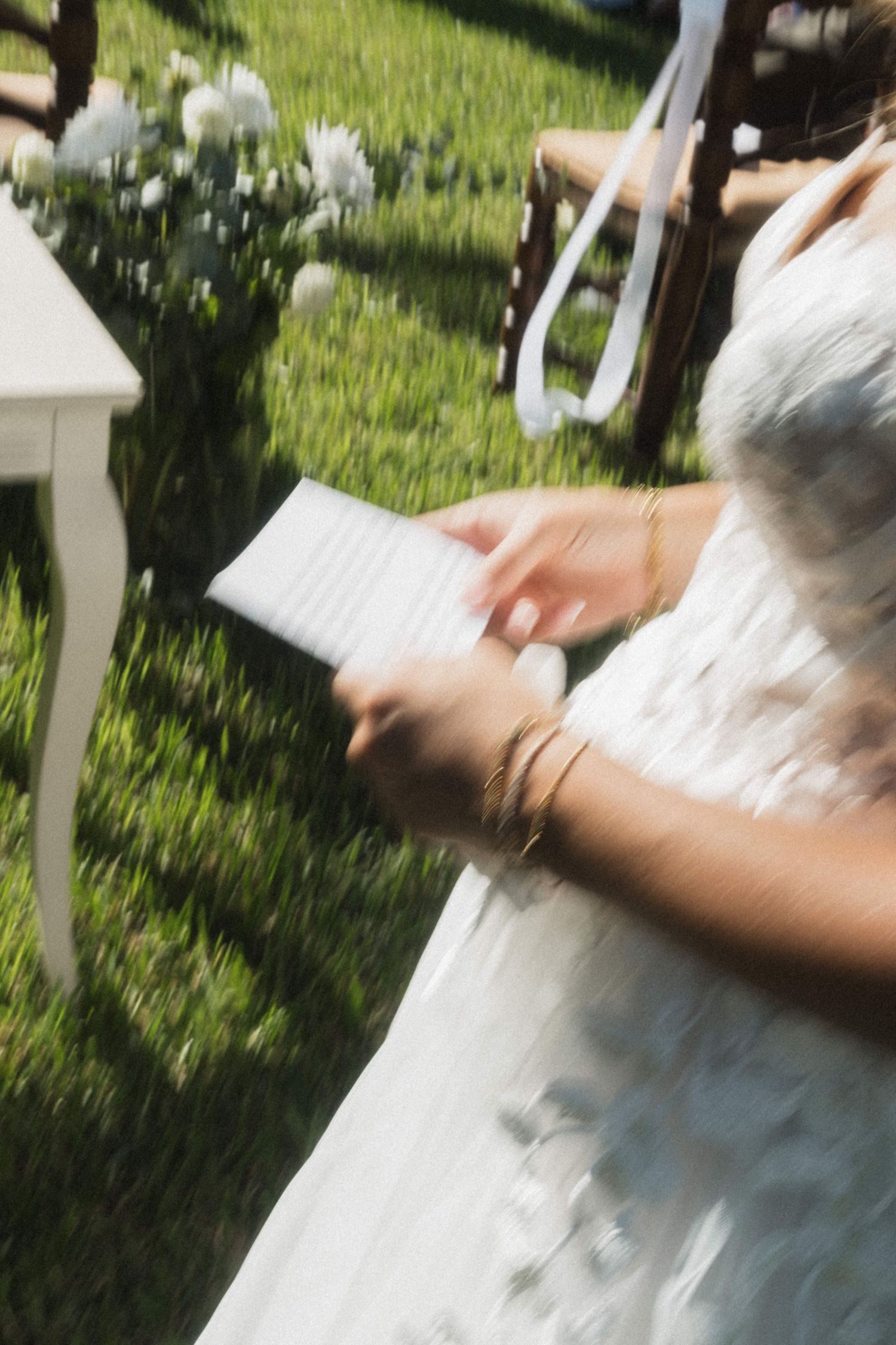Bride in white lace gown reading wedding vows from a card during outdoor ceremony.