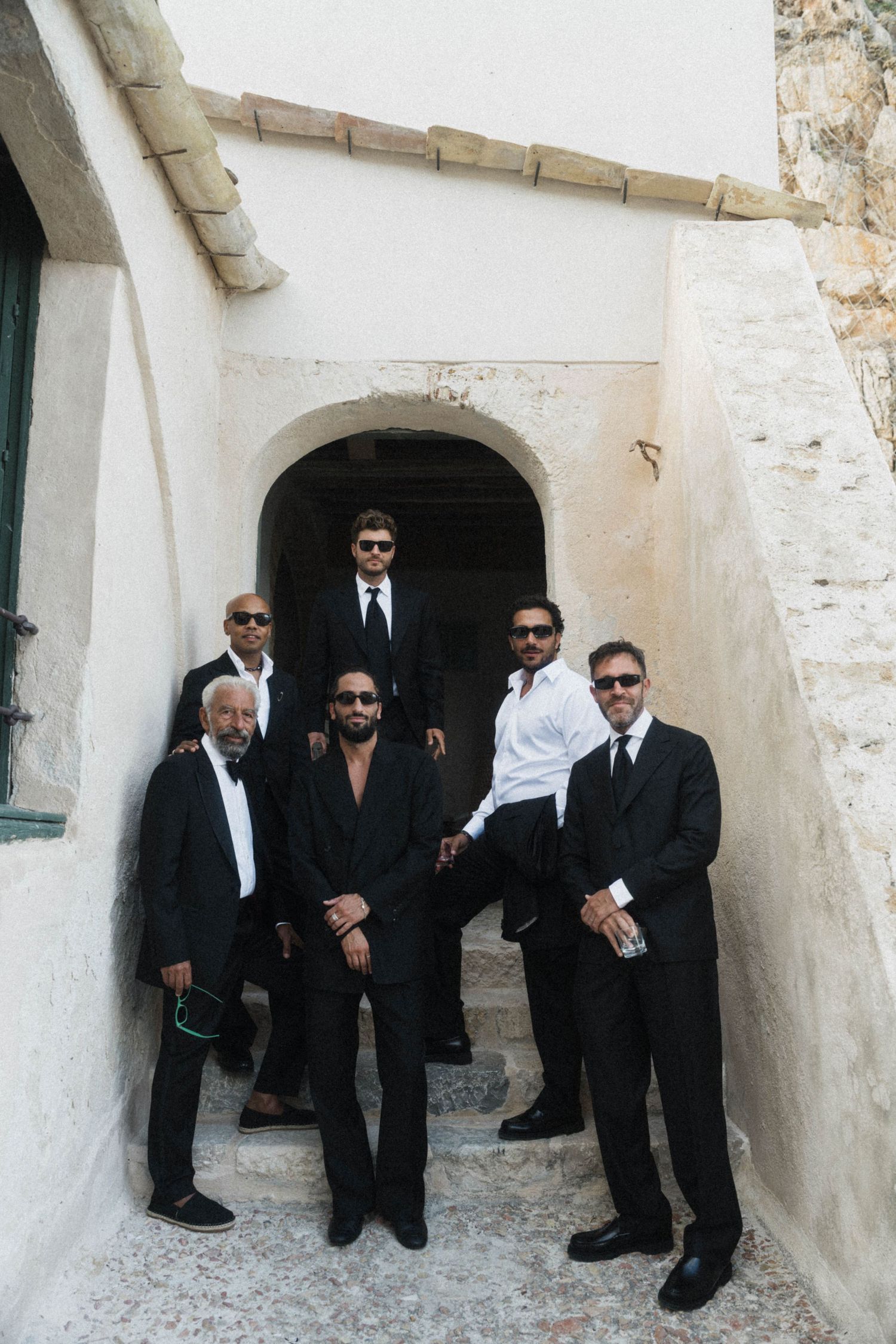 Group of six groomsmen in black suits posing on stone steps of a rustic building.