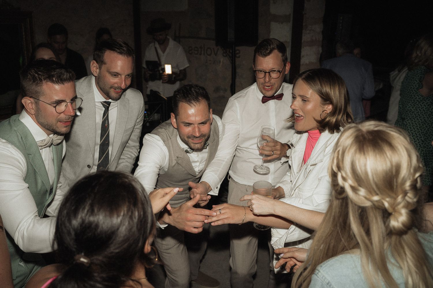 Group of elegantly dressed wedding guests clinking glasses in a joyful toast at an indoor reception.