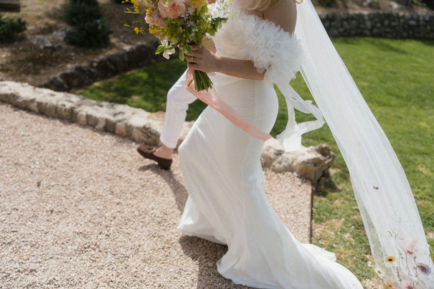 Bride in white gown and veil holding a floral bouquet walks along a stone path outdoors.