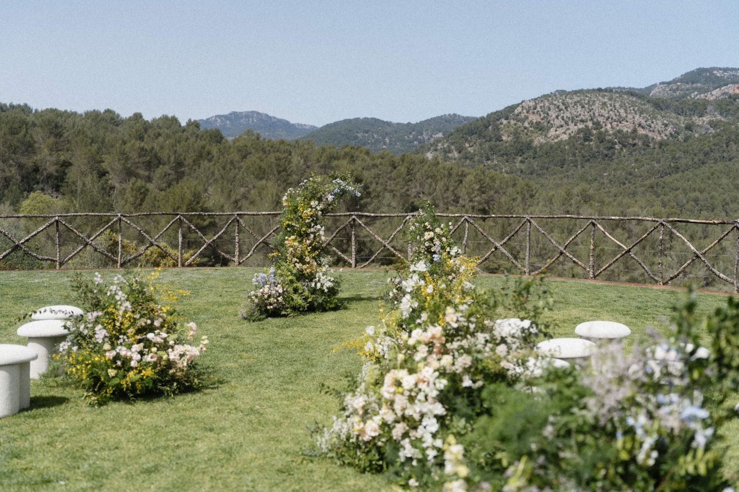 Outdoor wedding ceremony setup with floral arrangements on green lawn, rustic fence, and scenic mountain backdrop.