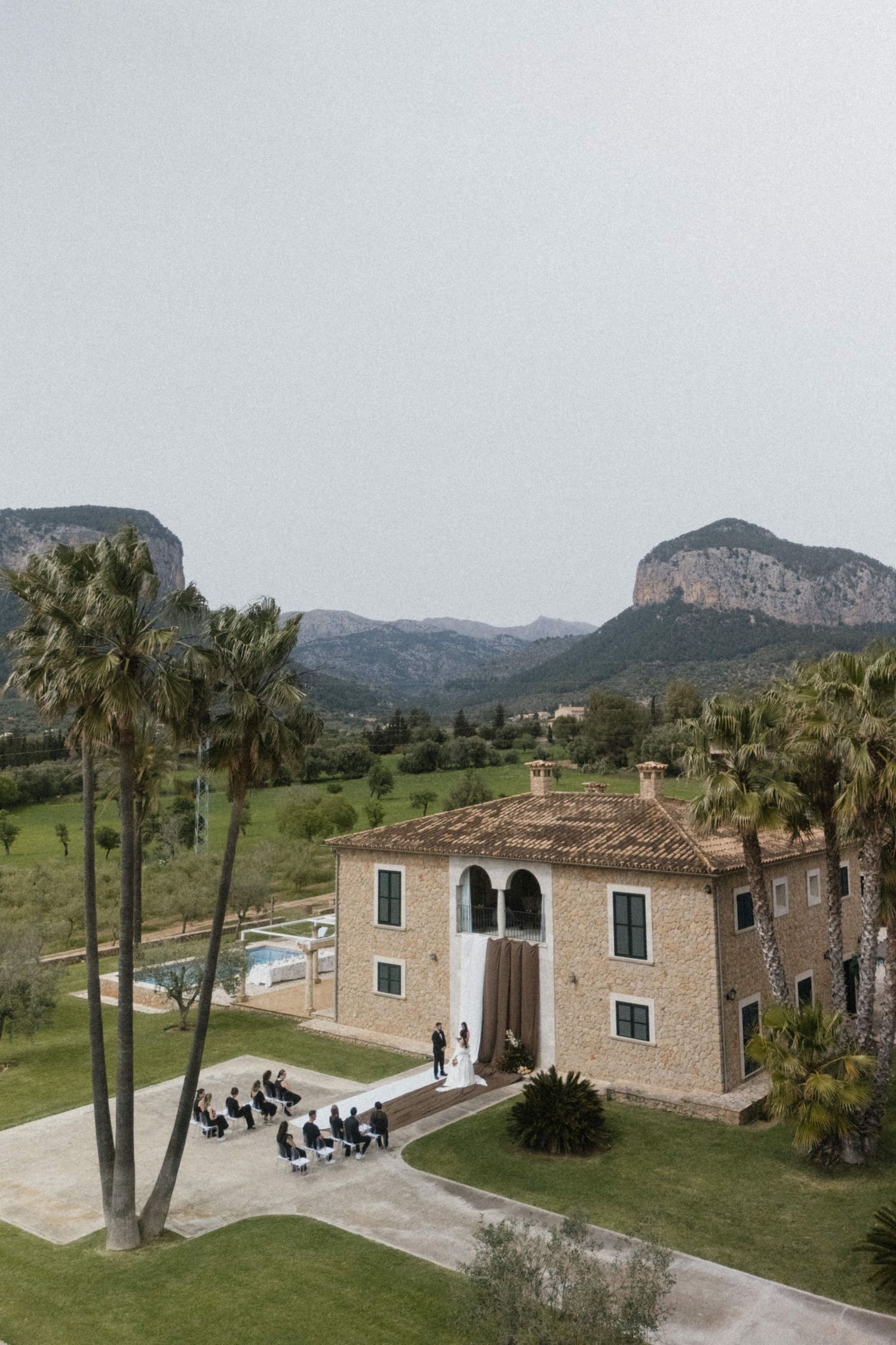 Aerial view of a rustic stone villa wedding venue surrounded by palm trees and mountains in Mallorca.