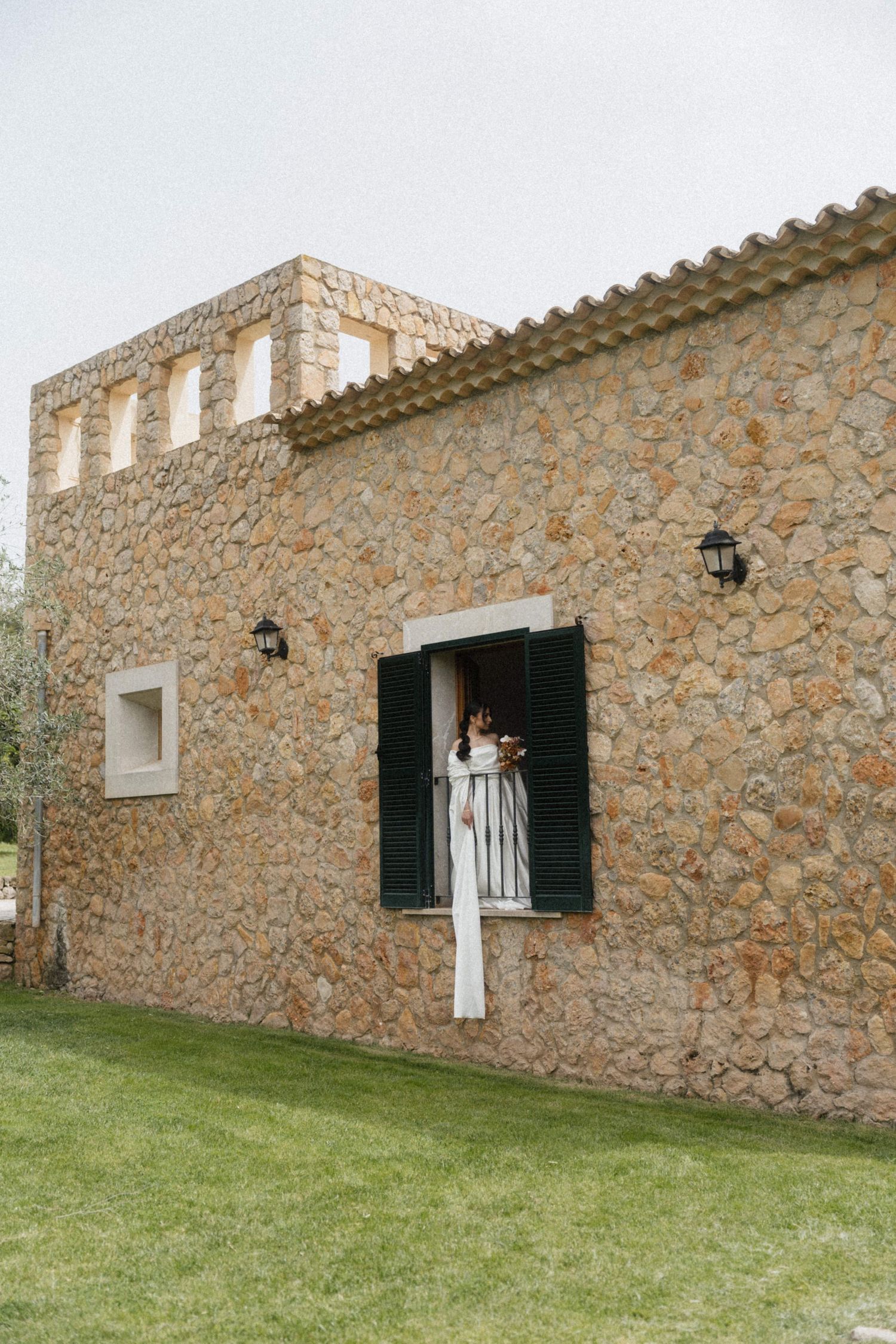 Bride in white gown standing on balcony of rustic stone villa with green shutters and terracotta roof.