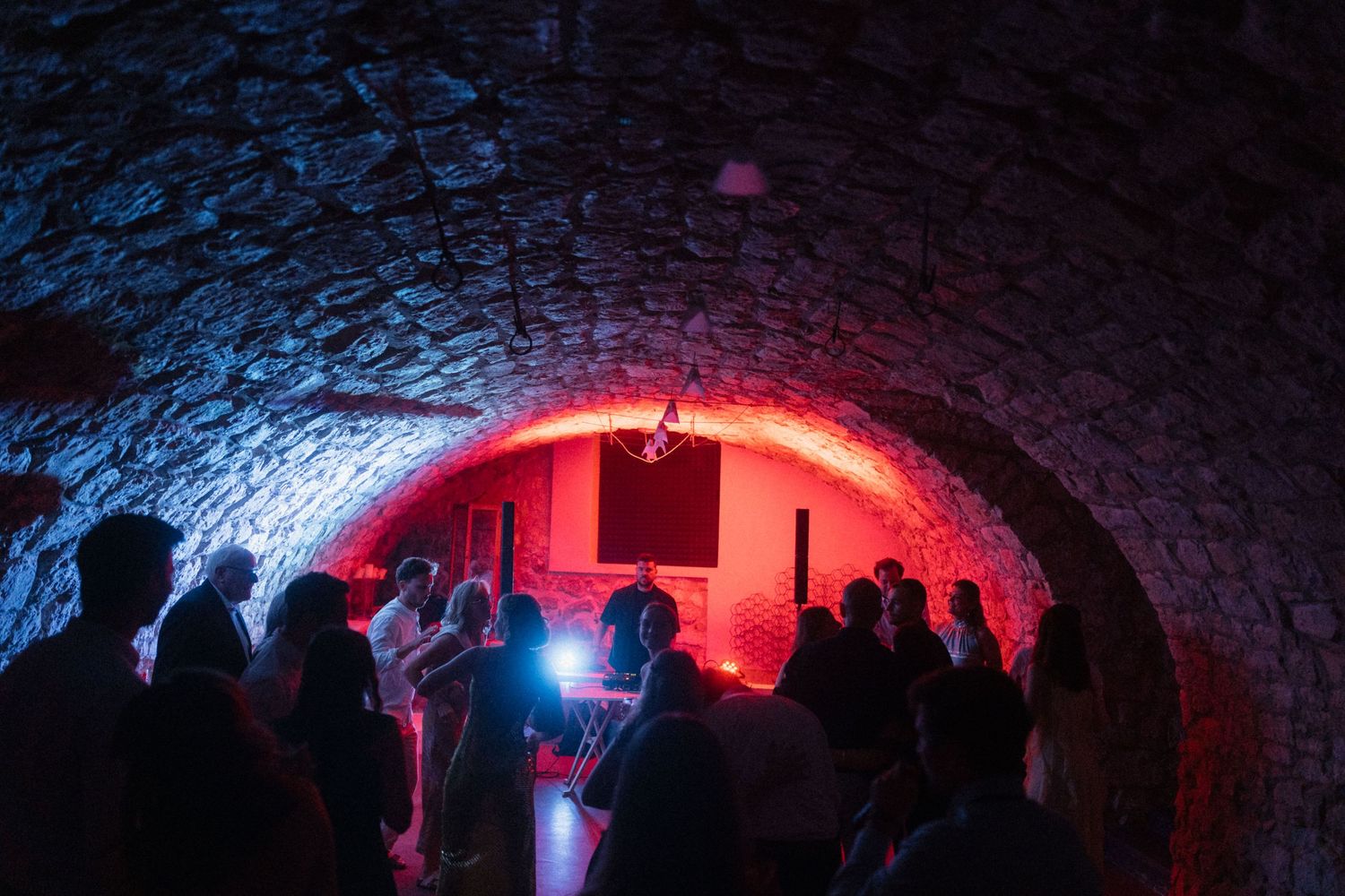 Wedding guests dancing in dramatic stone arch venue with red and blue atmospheric lighting at night.