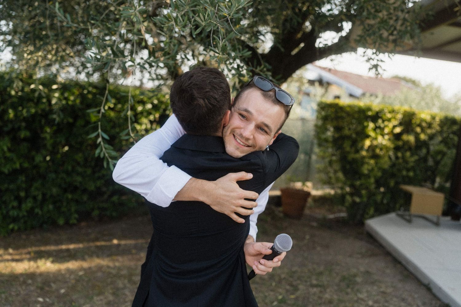 Two men sharing a warm hug outdoors at a wedding, one smiling broadly while holding a microphone.