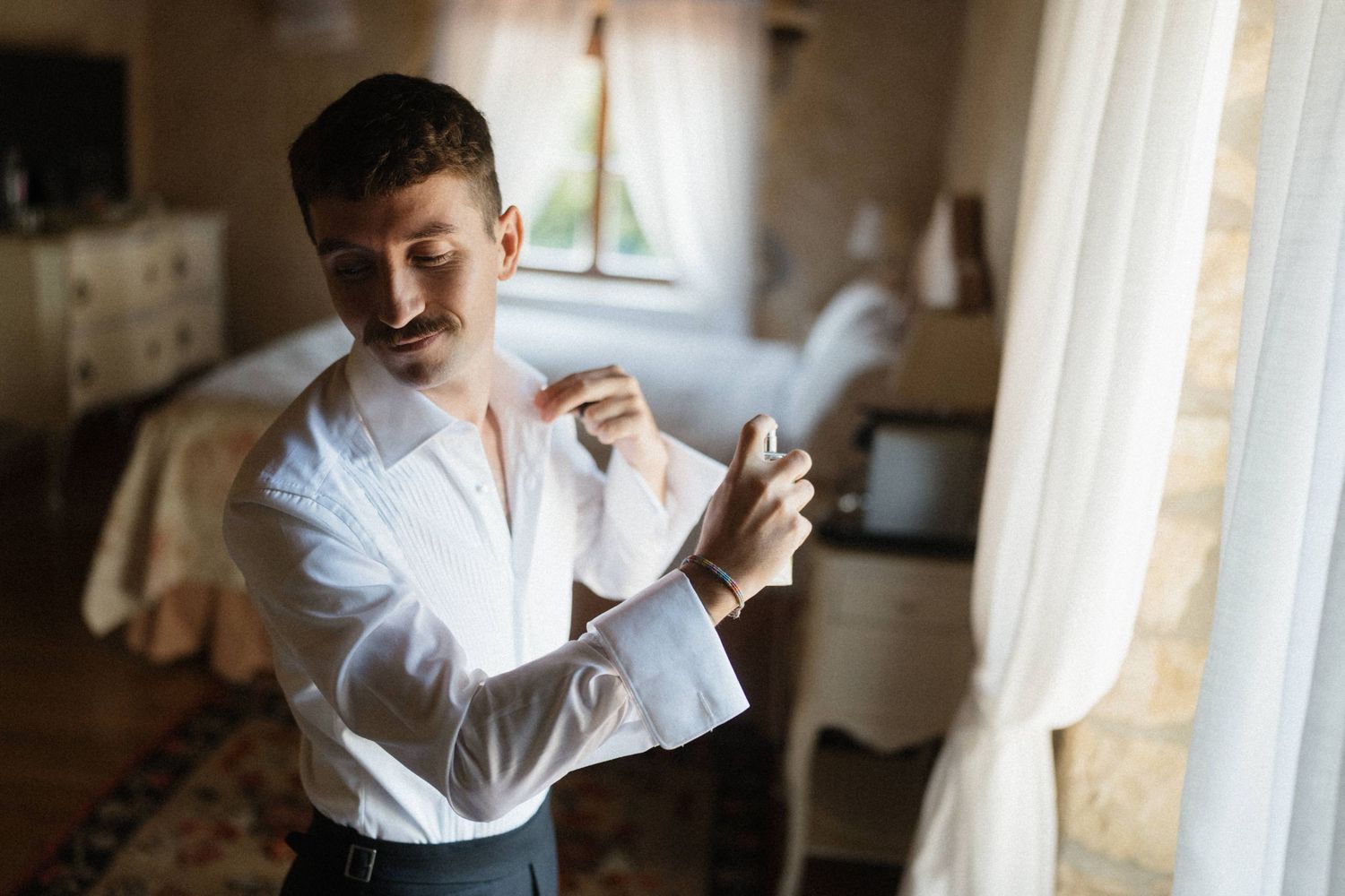 Groom in white dress shirt fastening cufflinks by a sunlit window in an elegant room.