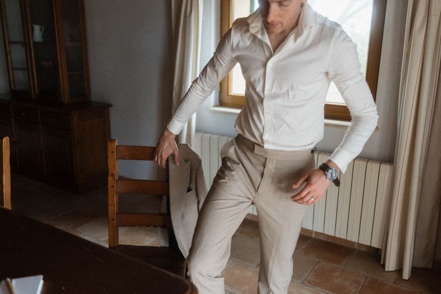 Man in white shirt and beige trousers standing by wooden chair in elegant room.