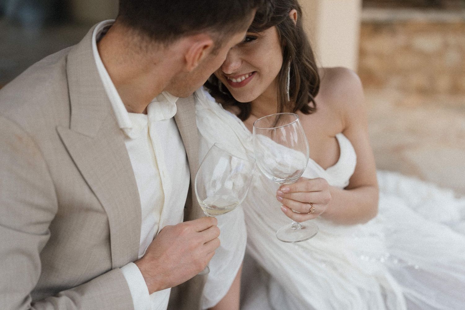 Smiling bride and groom in neutral wedding attire clink champagne glasses in a tender close-up moment.