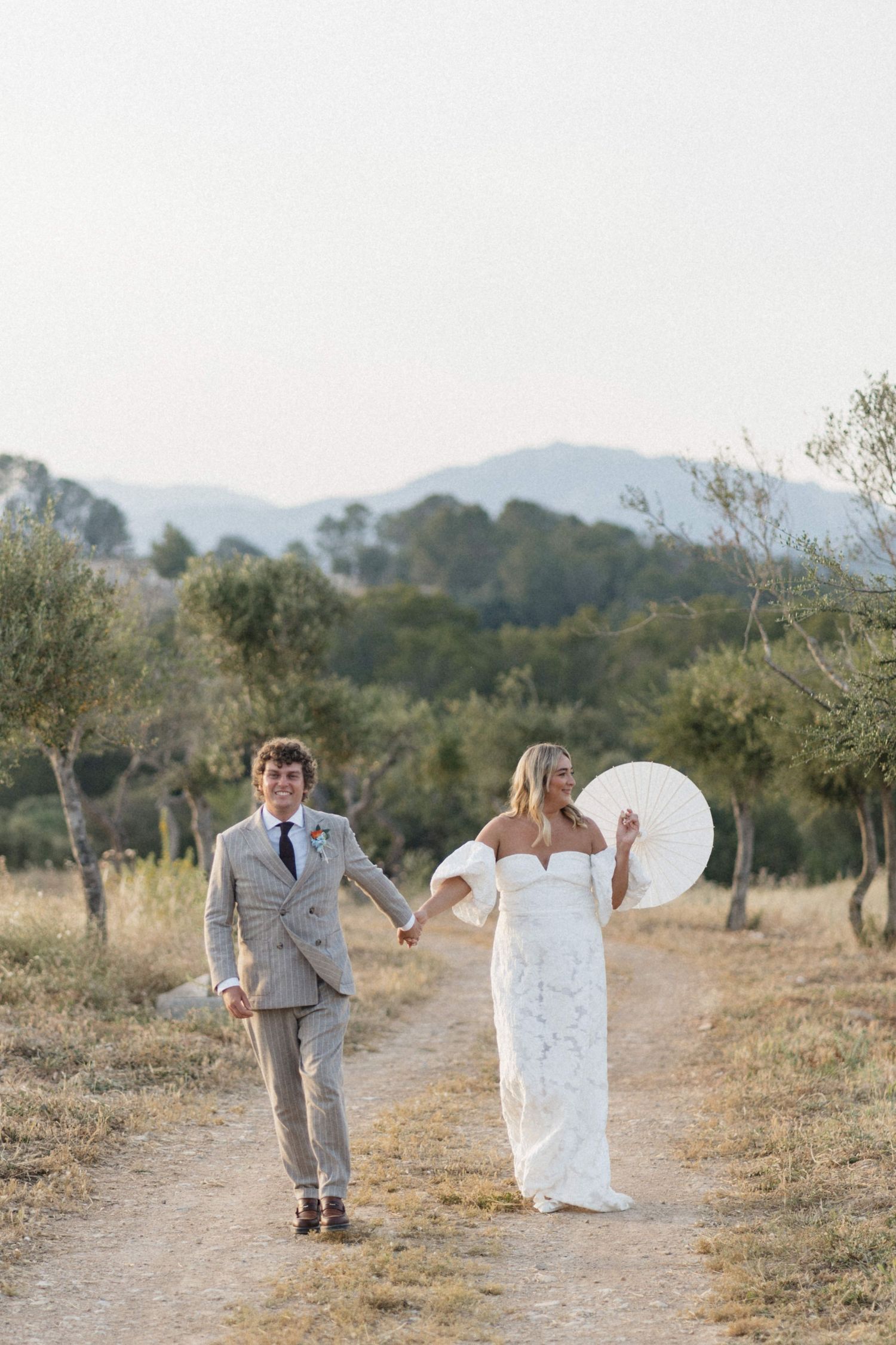 Newlywed couple walks hand-in-hand through an olive grove with scenic mountain backdrop on their wedding day.