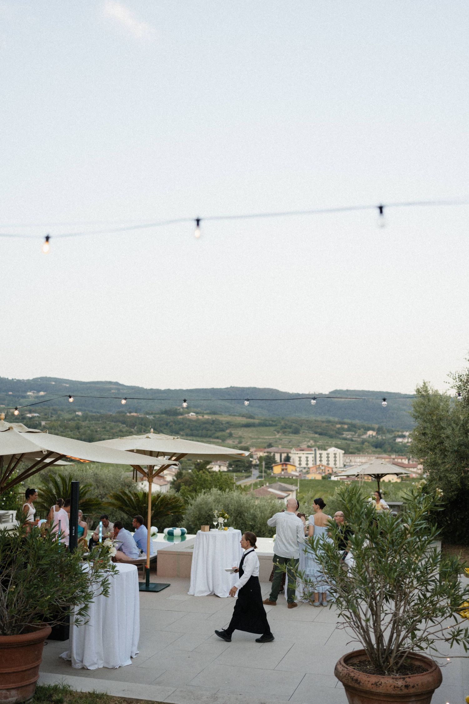 Outdoor wedding reception on a Tuscan terrace with string lights, white draped tables, and rolling hills at dusk.