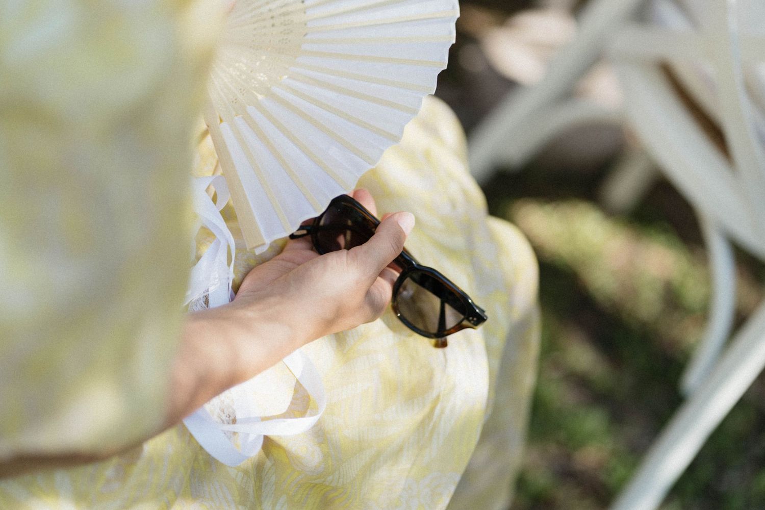 A hand holding sunglasses and a white paper fan at an outdoor wedding.