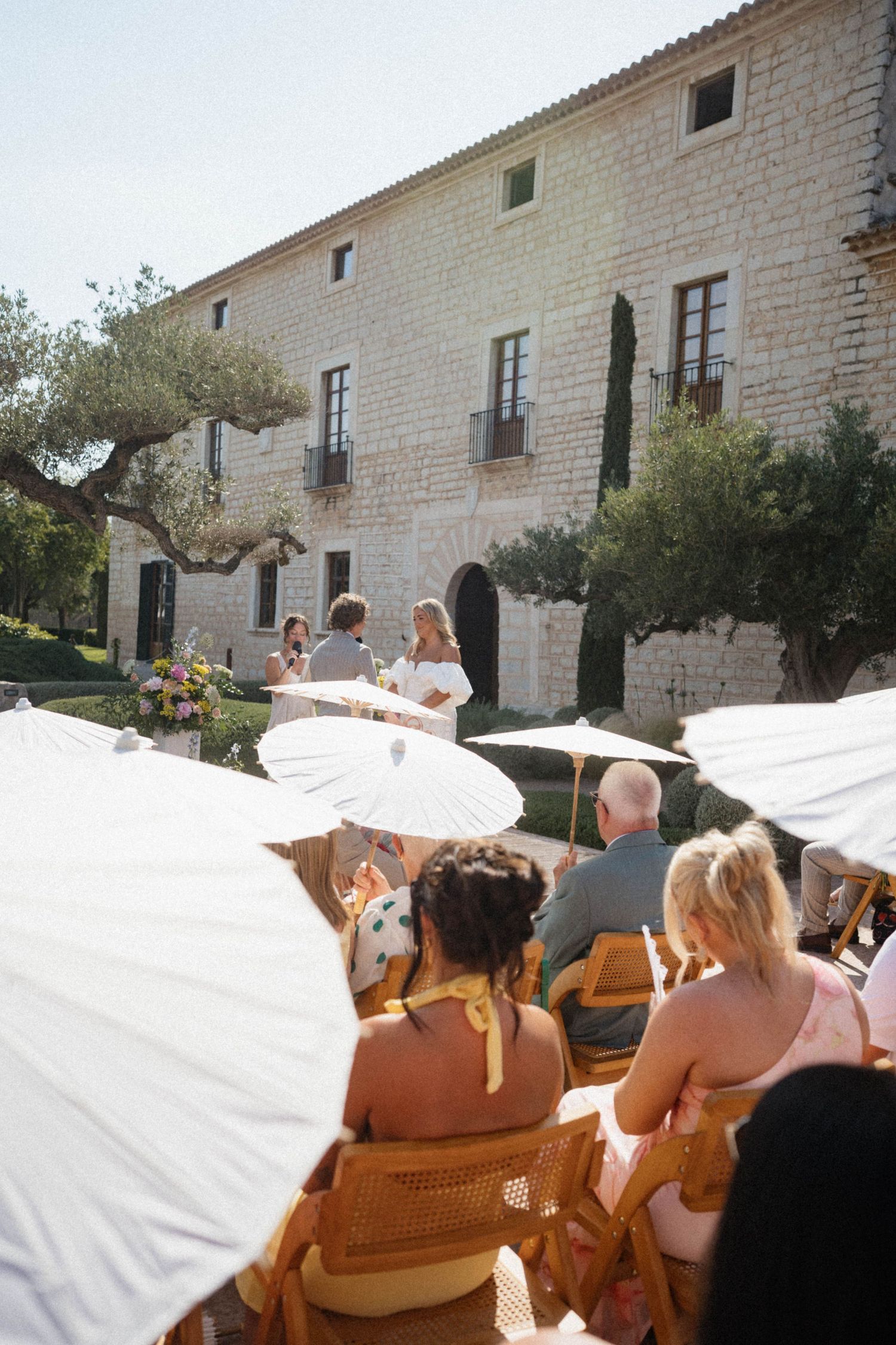 Wedding guests seated with white parasols watching outdoor ceremony at rustic stone villa venue.