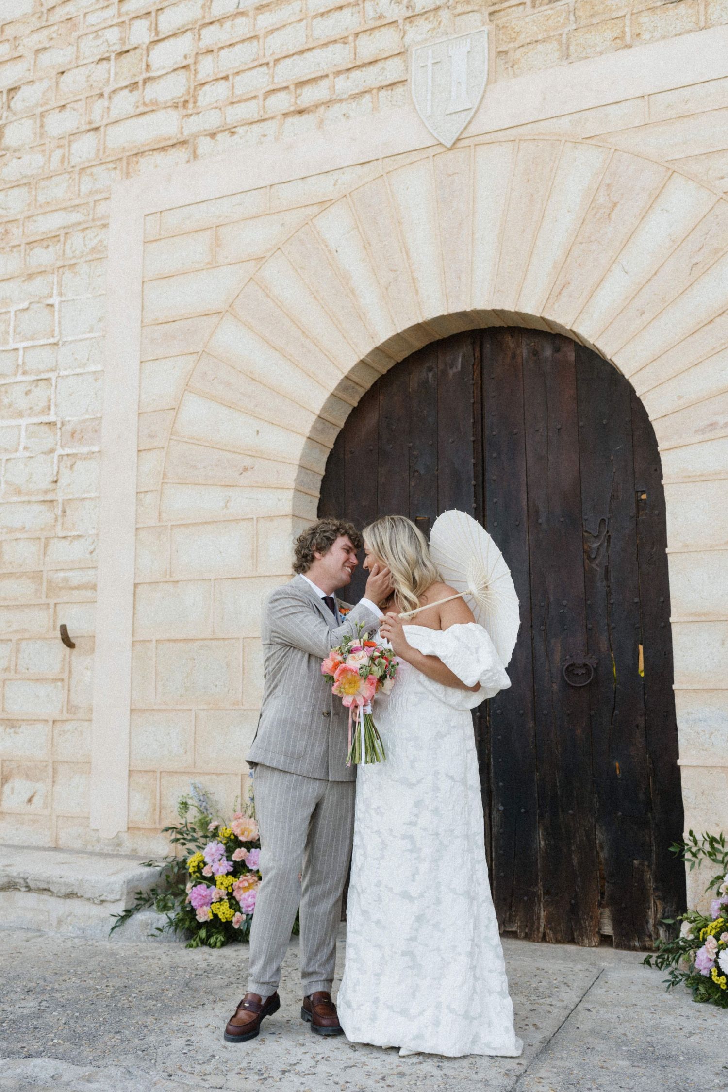 Bride and groom share a kiss in front of a rustic arched wooden door, bride holding a colorful bouquet in a white lace gown.
