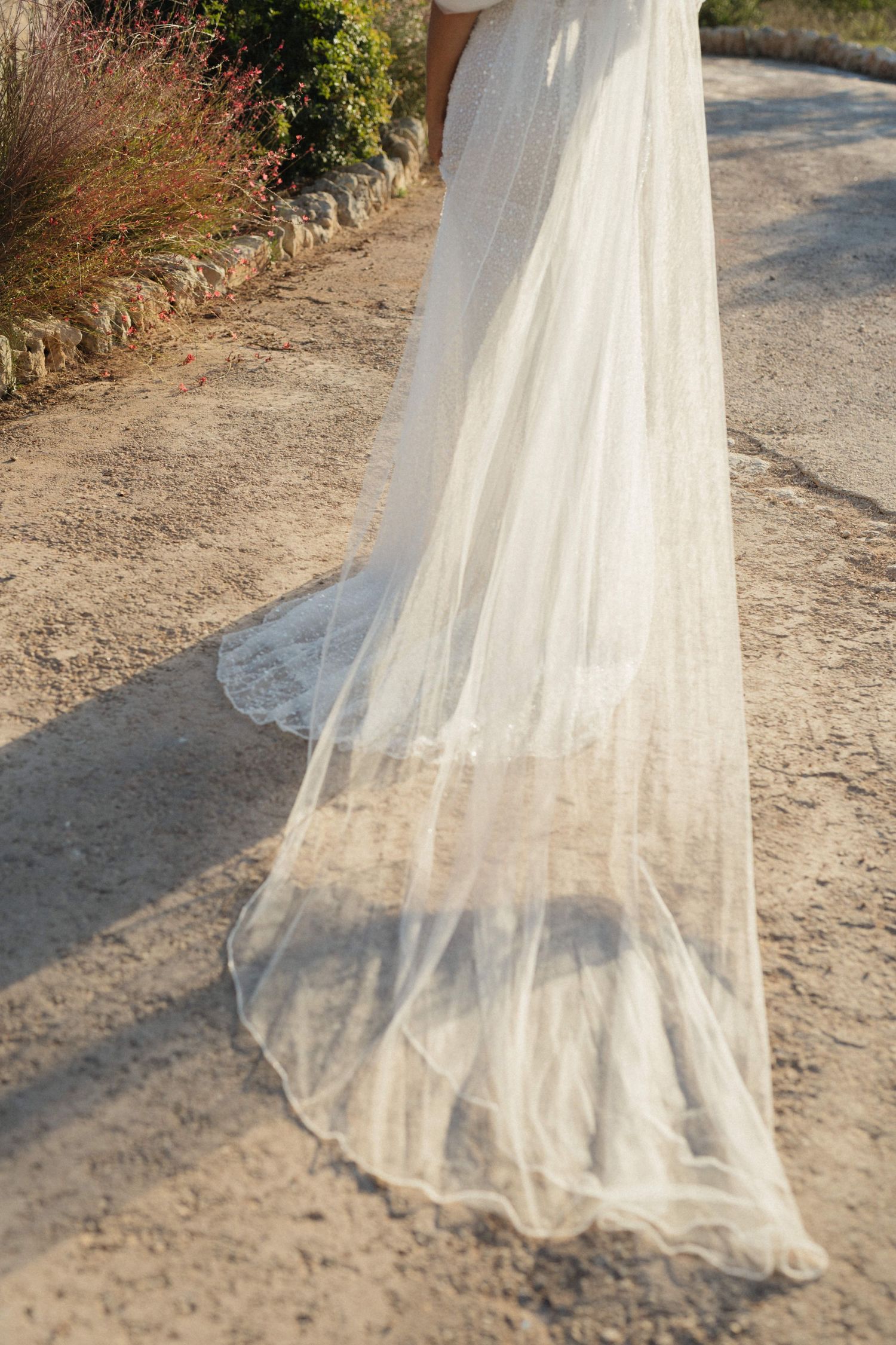 Long sheer bridal veil with delicate lace trim trailing over a sunlit gravel path at an outdoor wedding.