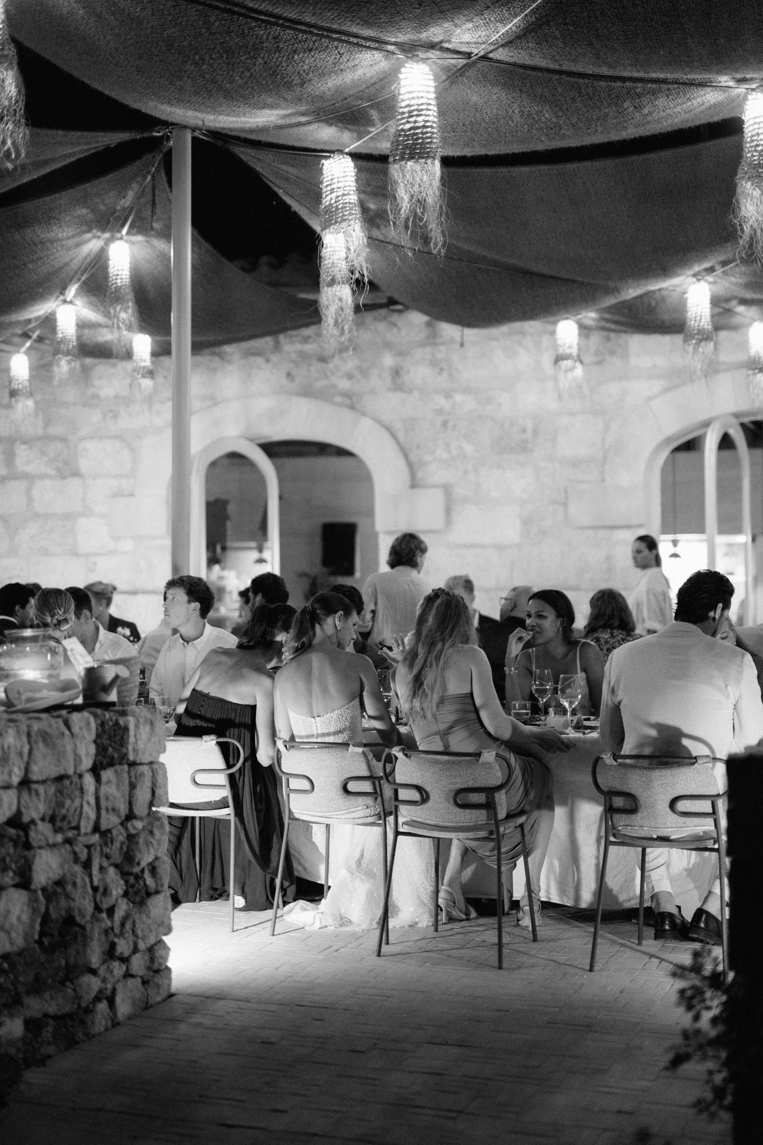 Black and white photo of wedding guests dining at a long table under a canopy with hanging lanterns beside a stone building.