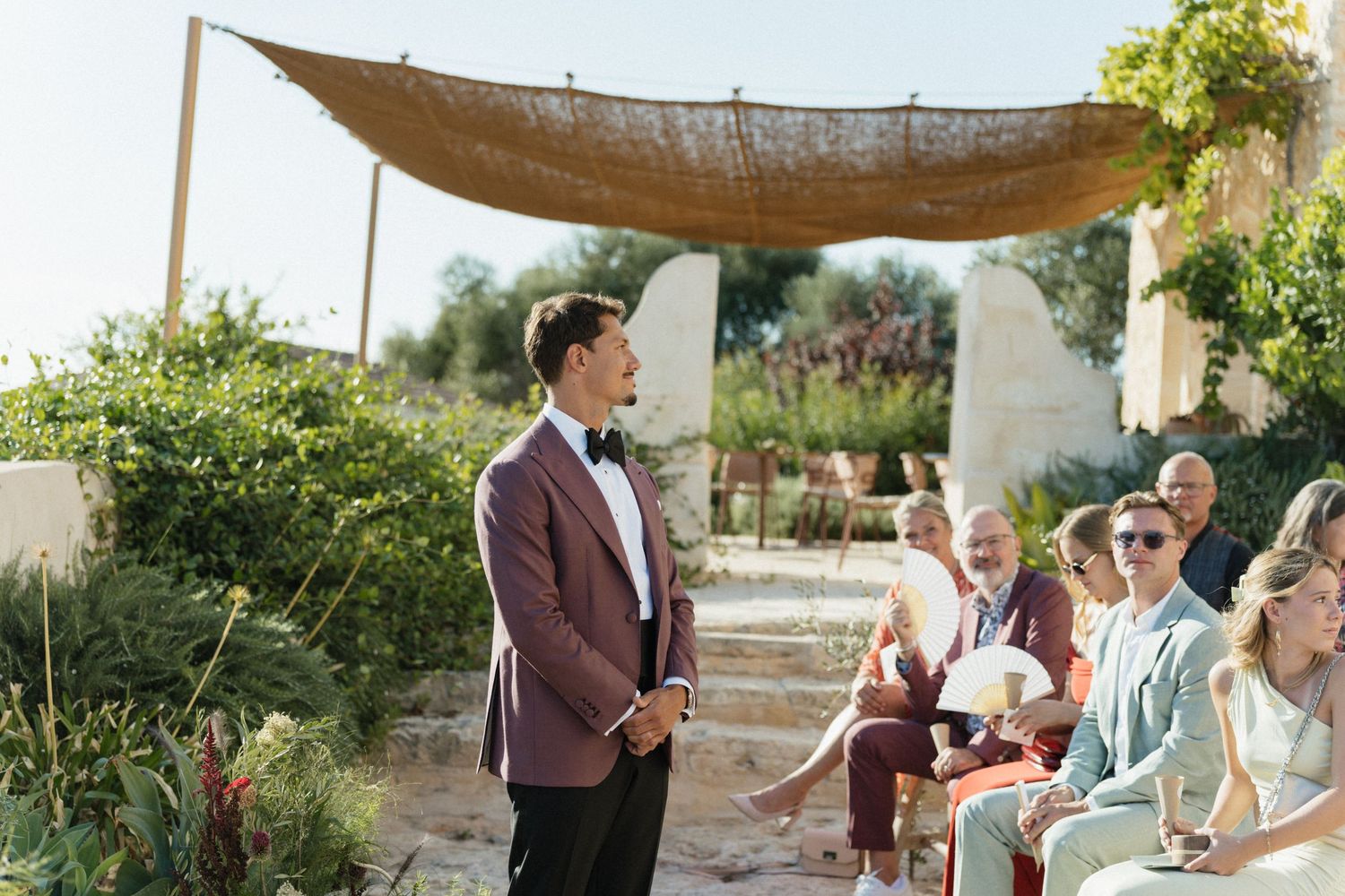 A groom in a burgundy jacket and bow tie waits at an outdoor wedding ceremony as guests sit under a woven canopy.