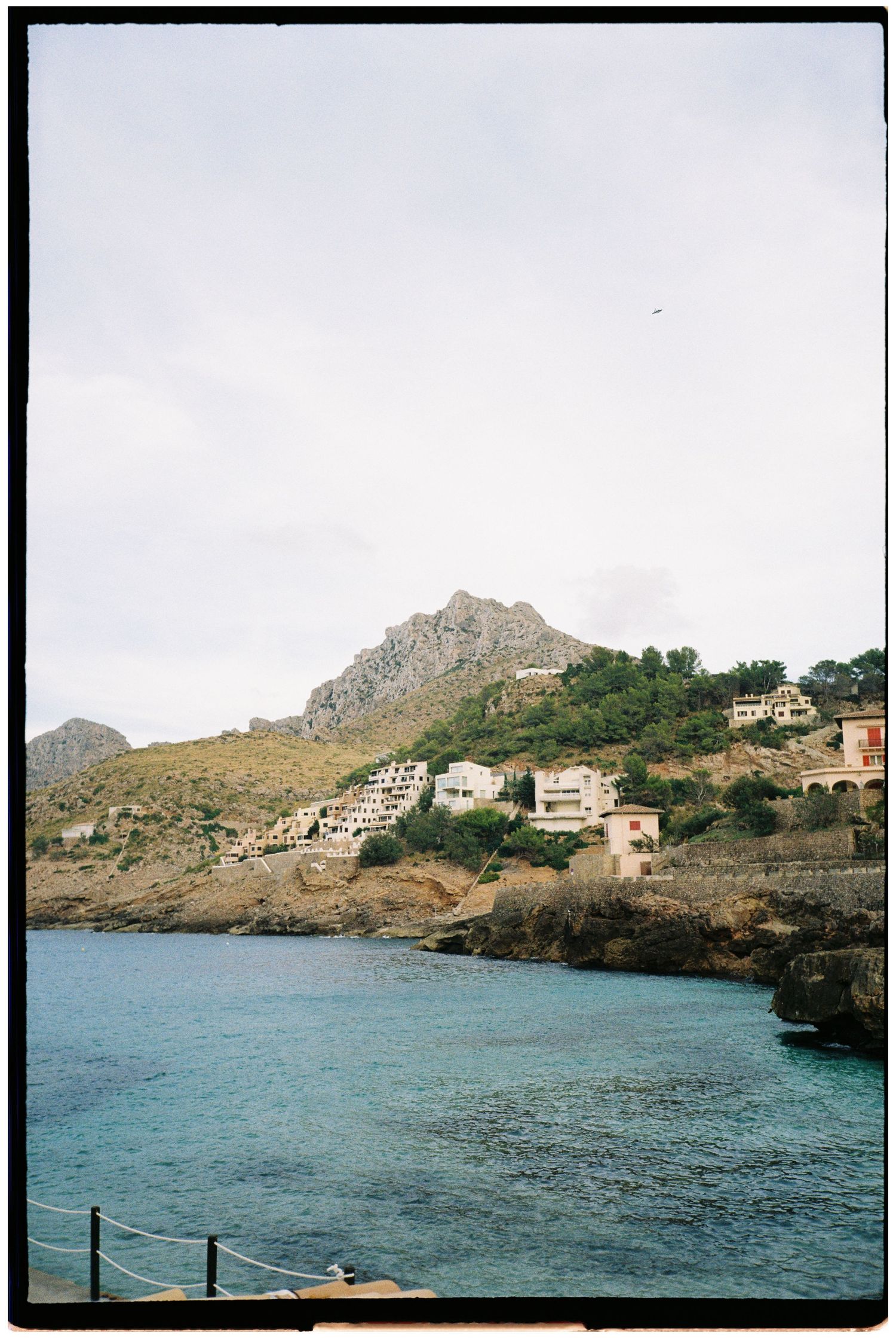 Whitewashed hillside villas overlook a calm turquoise bay beneath a rugged mountain peak on a cloudy day along the Mediterranean coast.