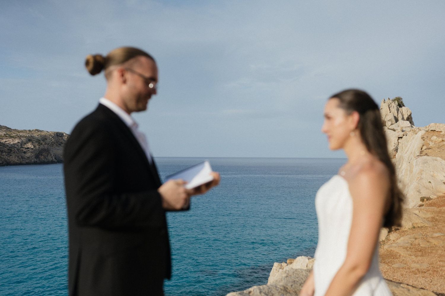 Groom in black suit reads vows from a small book to a bride in a white dress during a seaside elopement on rocky cliffs above the blue sea.