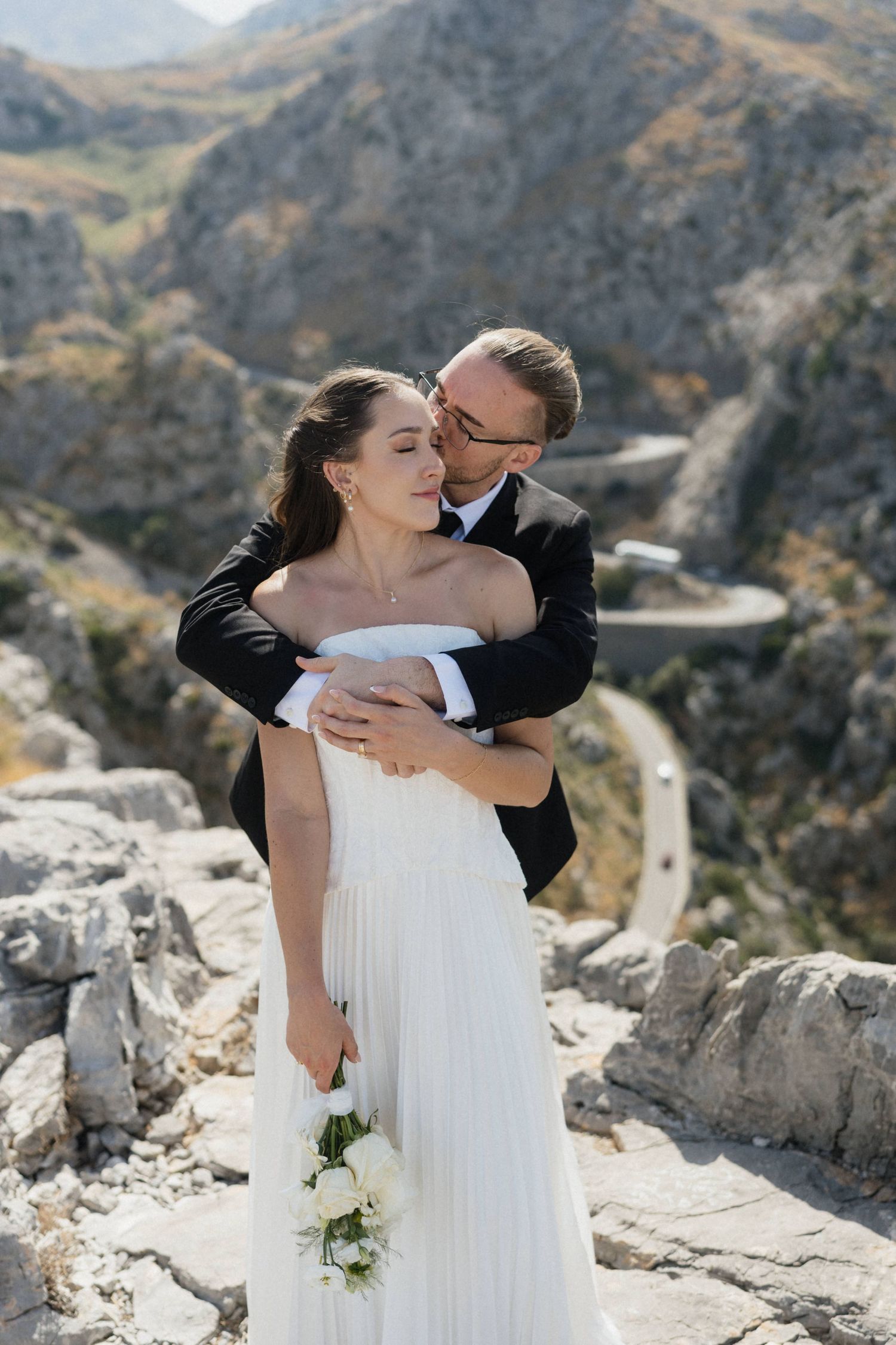 Groom in a black suit embraces a bride in a white pleated gown holding a bouquet on a rocky mountain overlook with winding roads.