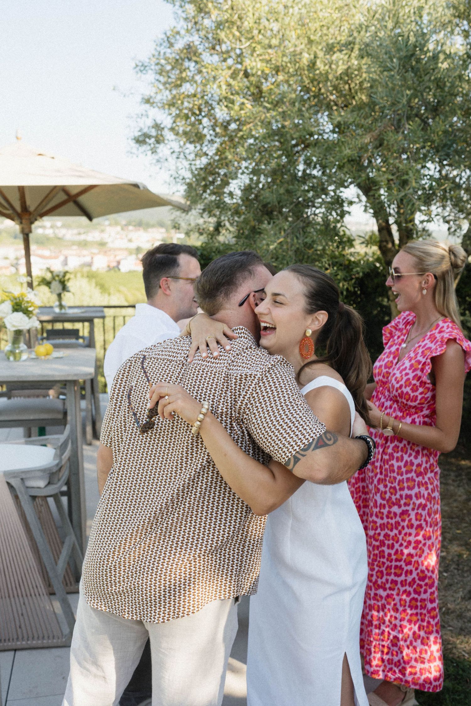 Guests warmly embracing at an outdoor summer party with trees and scenic views in the background.