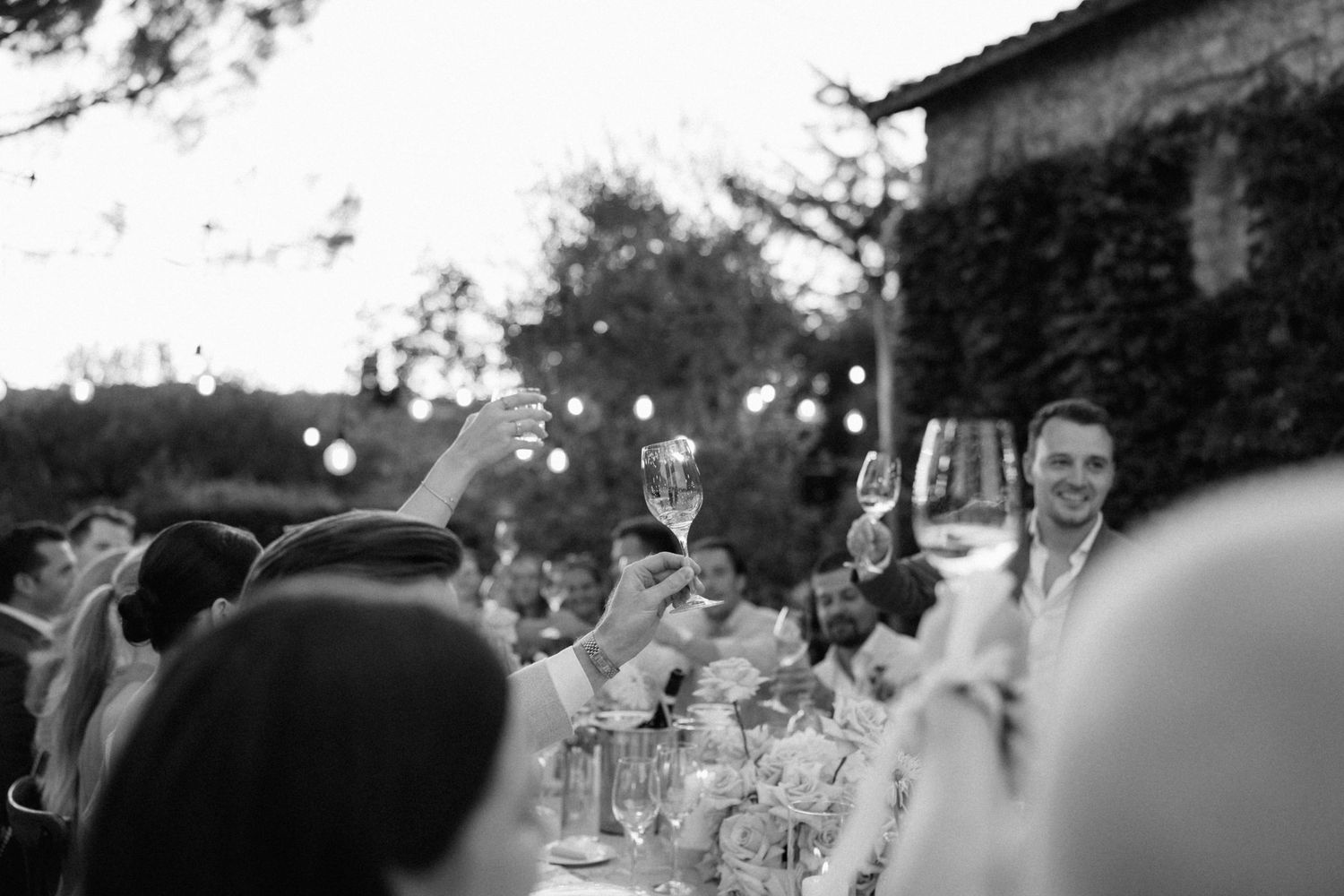 Black and white photo of wedding guests raising glasses in a toast at an outdoor reception with string lights.