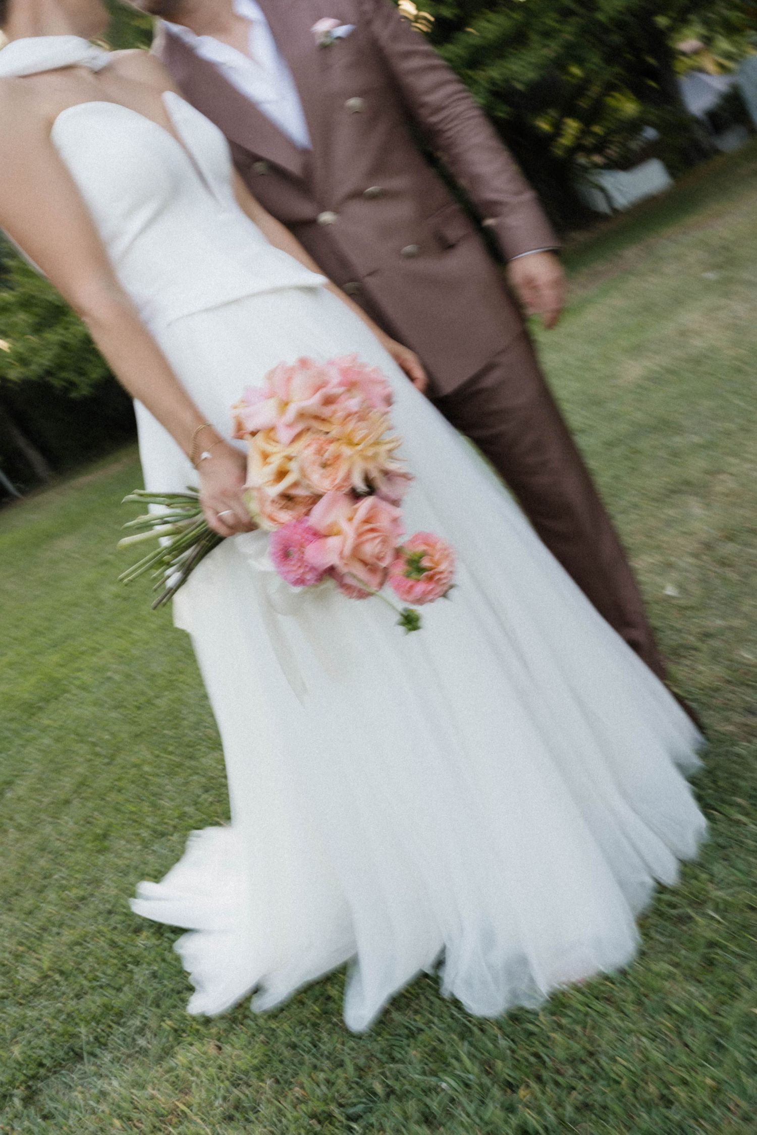 Bride in white tulle gown holding pink floral bouquet beside groom in brown suit outdoors.