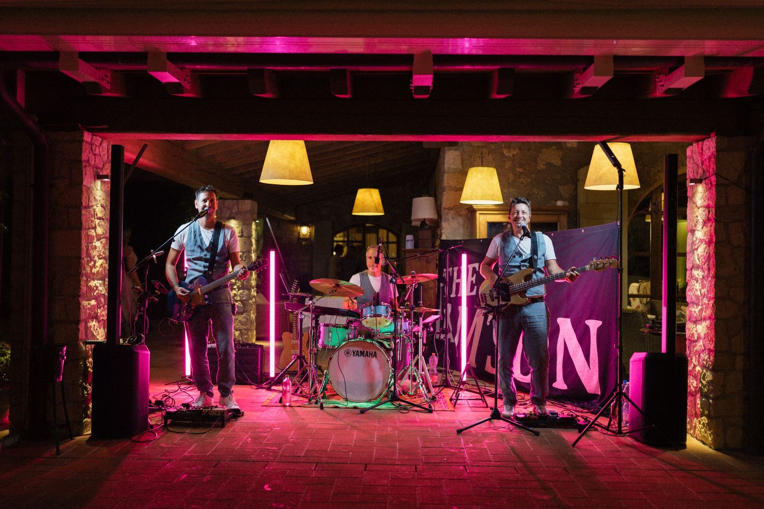 A live band performs on a pink-lit stage with guitars and drums at an indoor venue.
