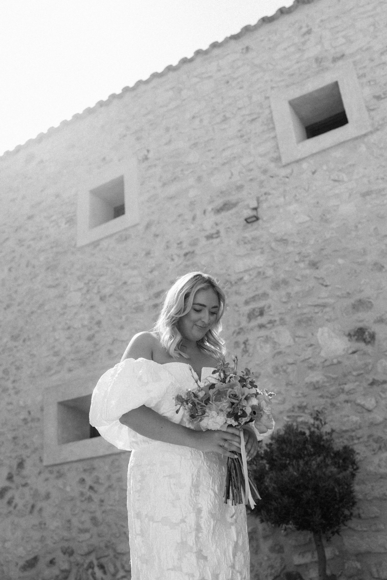 Black and white portrait of bride in lace gown holding bouquet outside rustic stone building.
