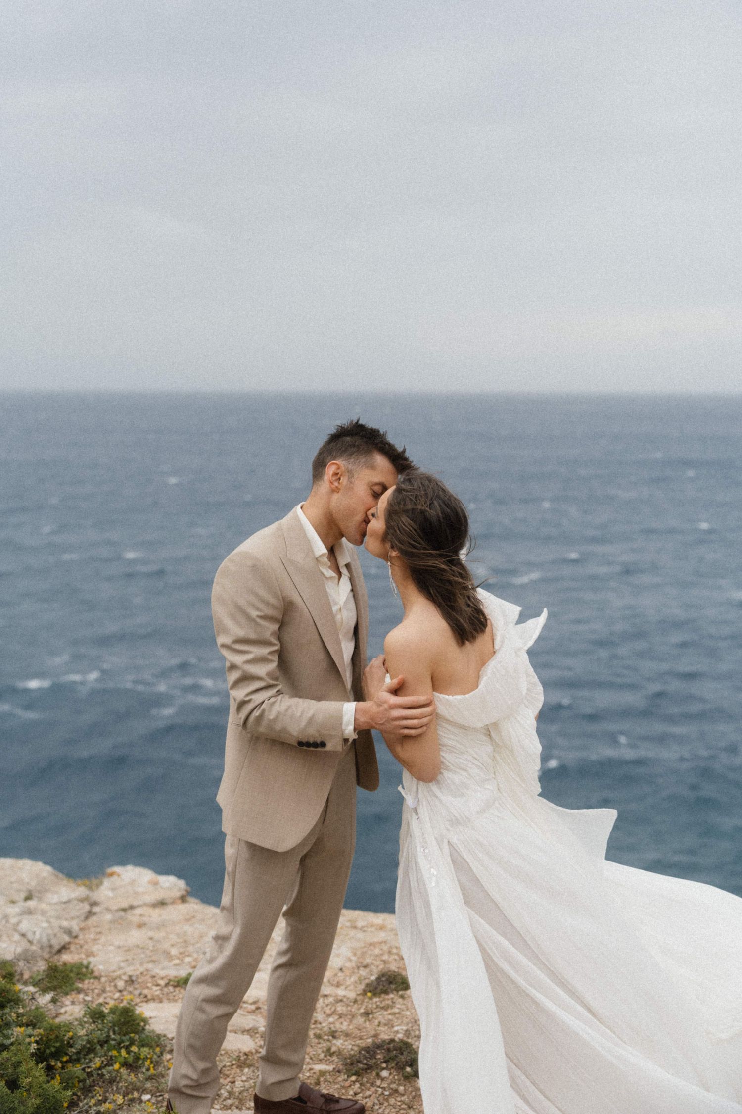 Bride and groom sharing a romantic kiss on a rocky cliff overlooking the deep blue sea.