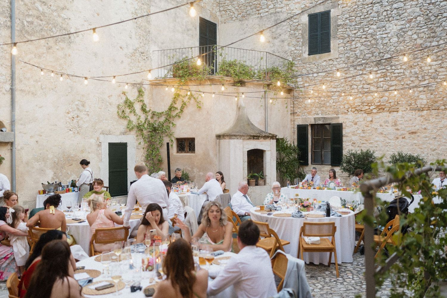 Outdoor wedding reception in a rustic stone courtyard with string lights, guests dining at elegant tables.