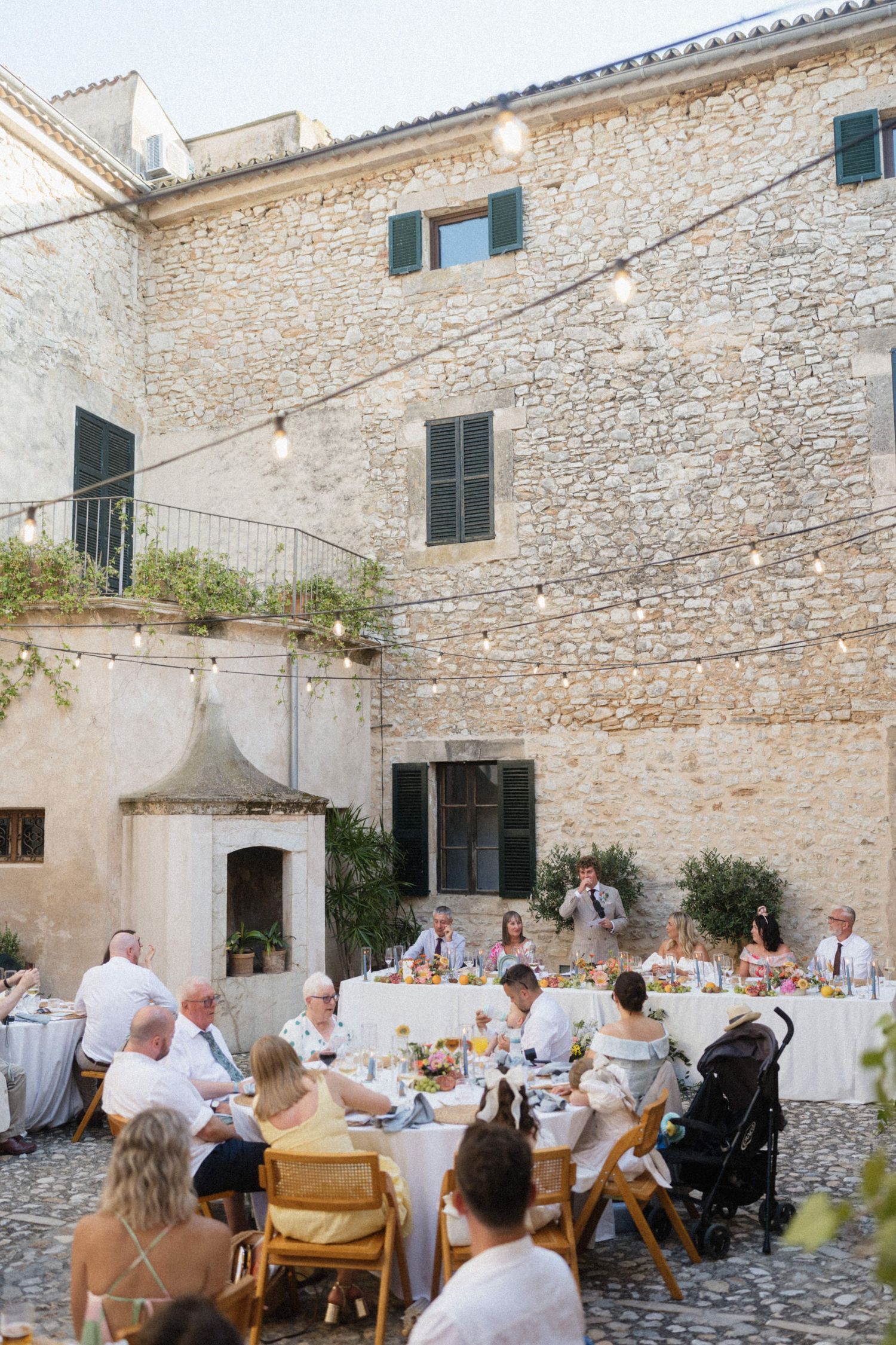 Wedding guests dine al fresco in a charming cobblestone courtyard of a historic stone building with string lights.