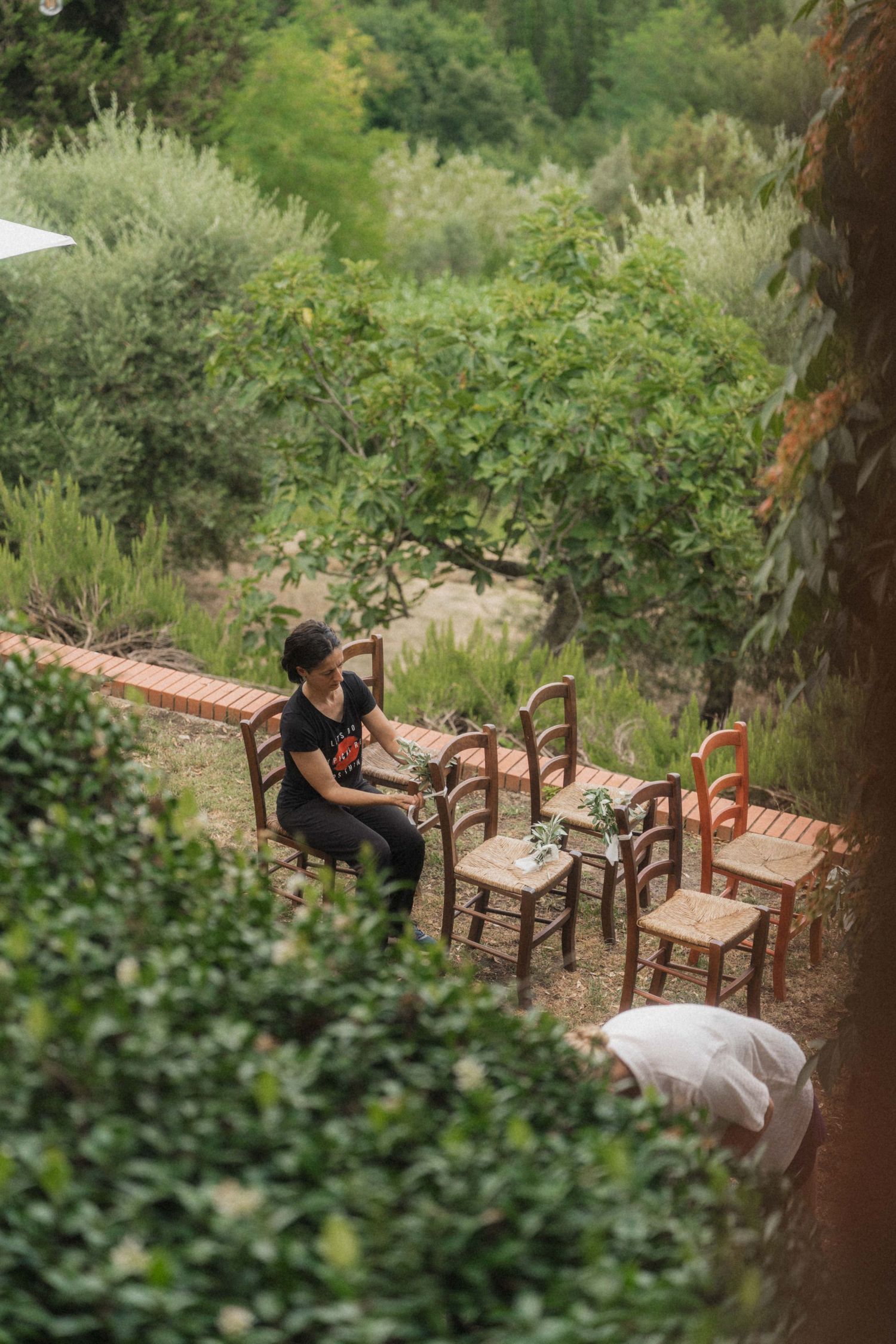 Woman arranging wooden chairs at an outdoor garden wedding venue surrounded by lush greenery.