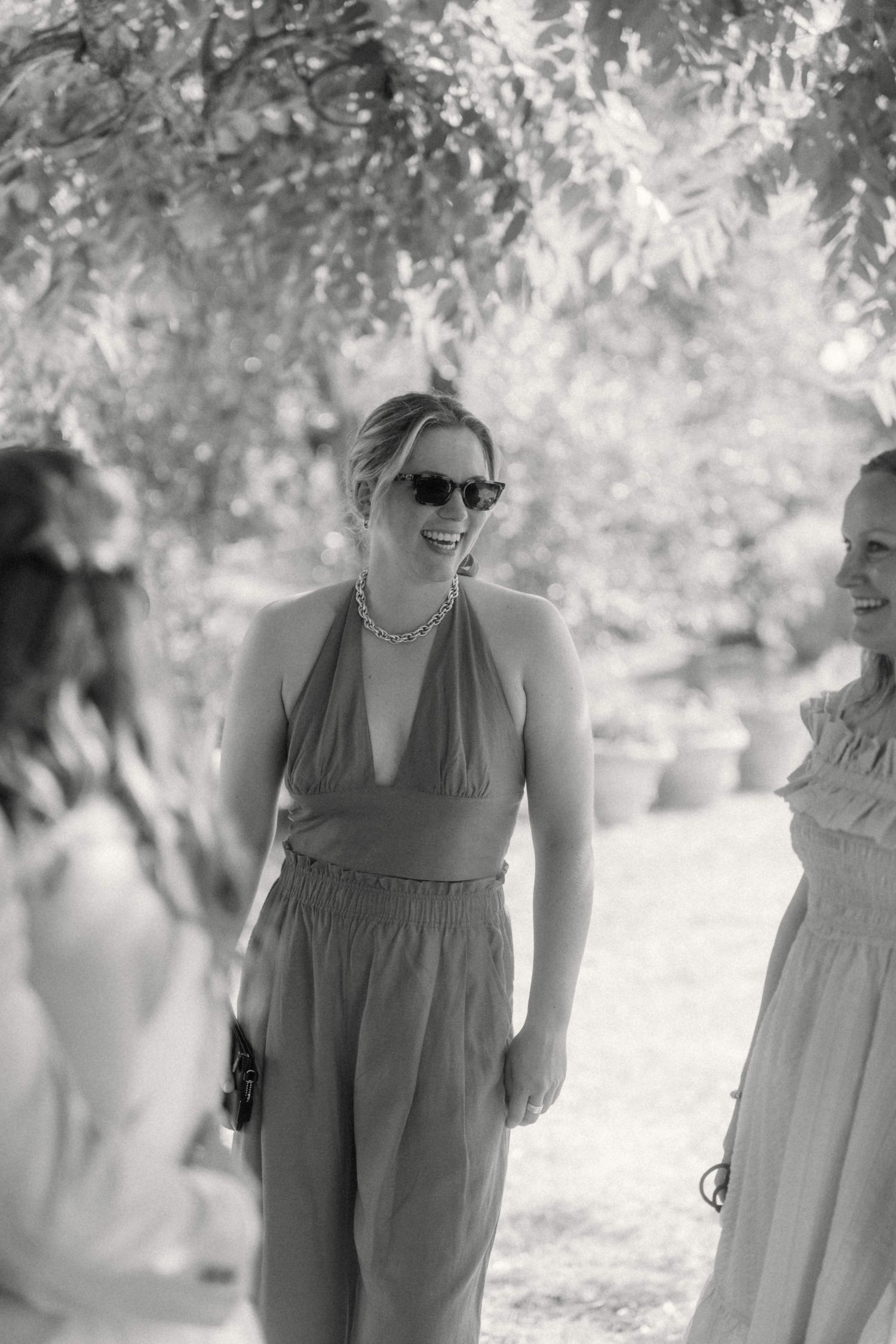 Black and white photo of smiling wedding guest in halter jumpsuit and sunglasses at outdoor celebration.