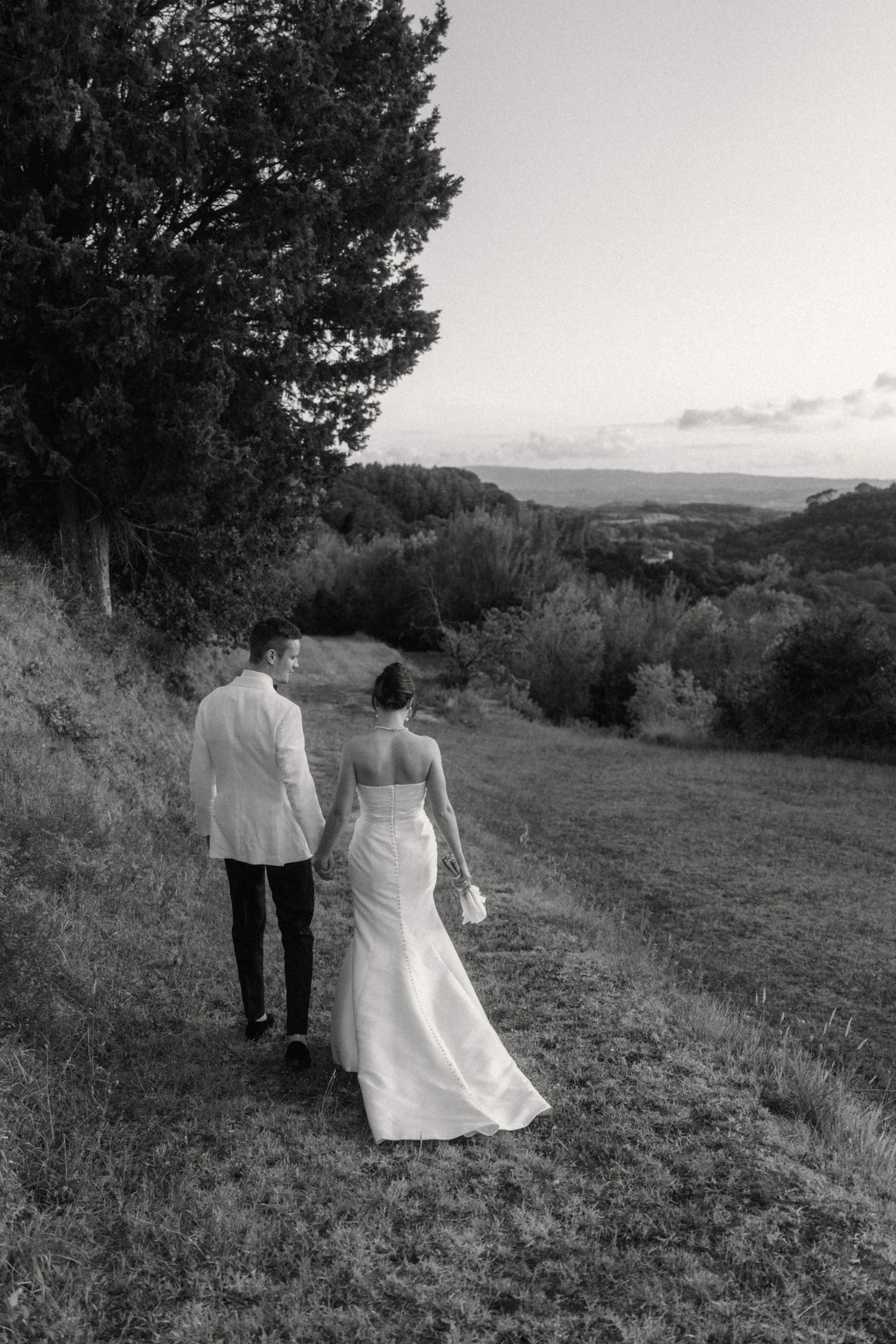 Black and white photo of bride and groom walking hand in hand through Tuscan countryside at dusk.