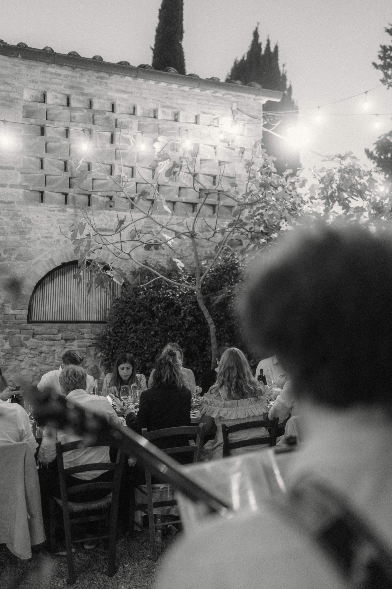 Black and white photo of wedding guests dining outdoors against a rustic stone wall with string lights at dusk.
