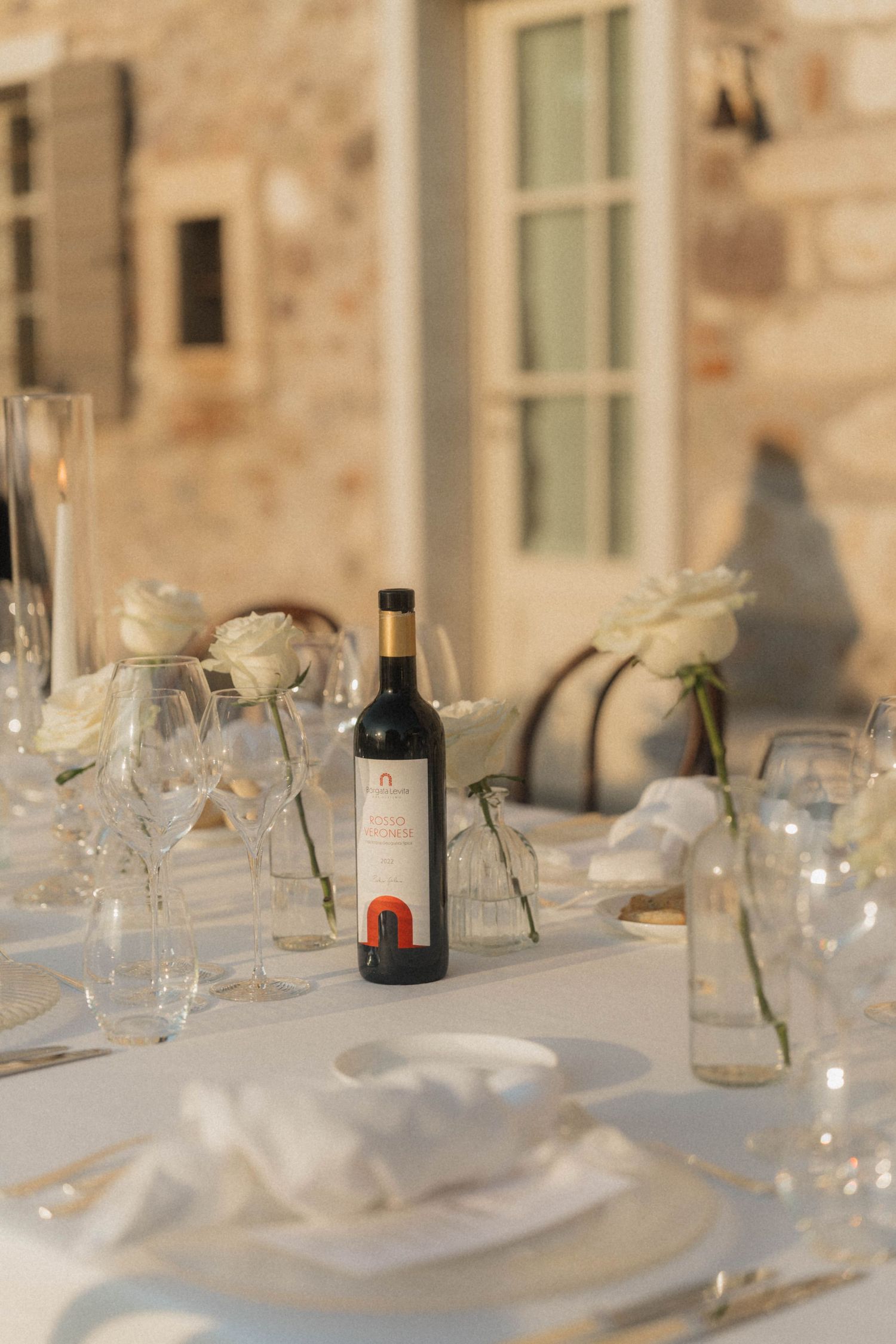 Elegant outdoor wedding reception table set with wine bottle, white roses, crystal glasses, and linen napkins.