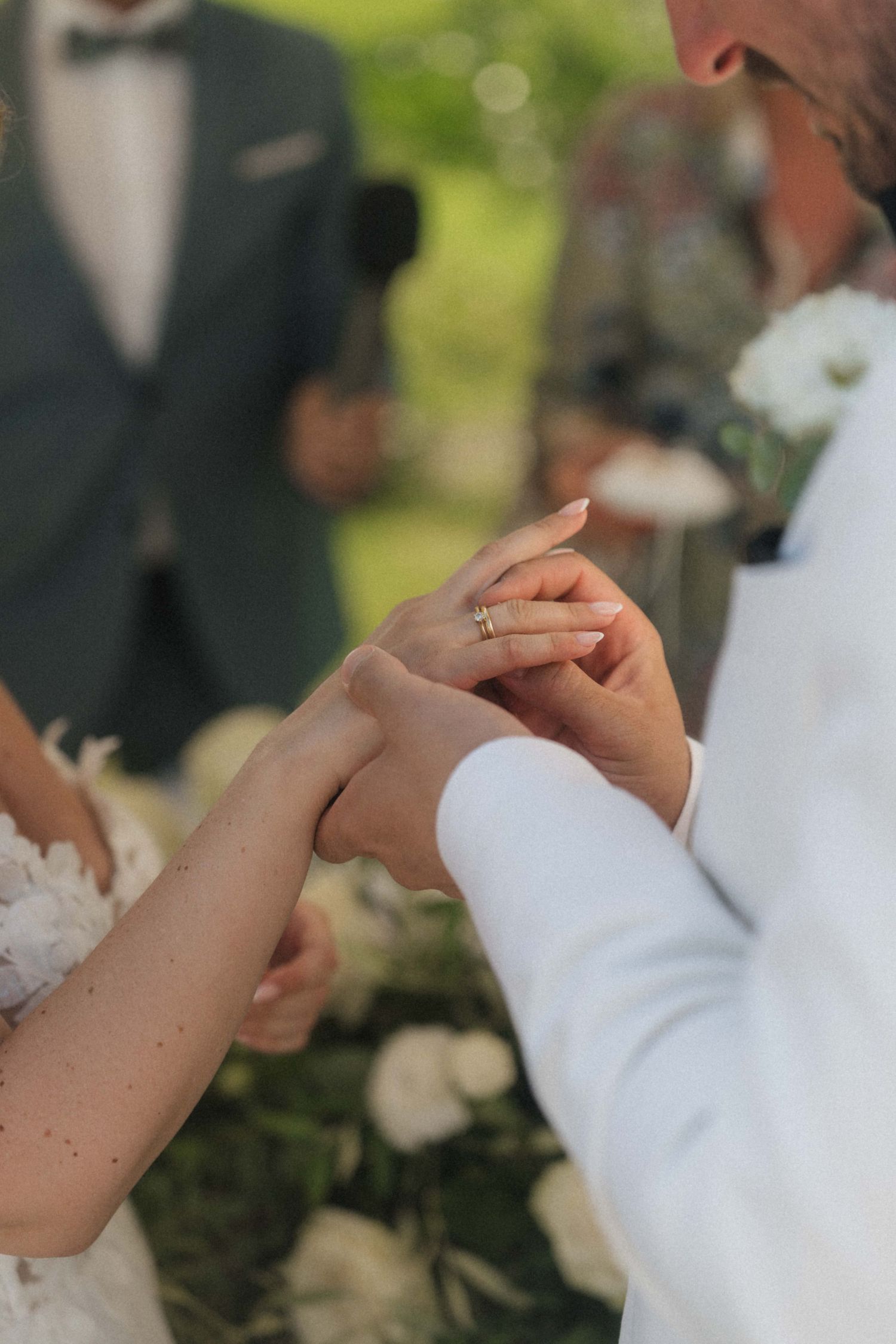 Groom places wedding ring on brides finger during outdoor wedding ceremony with white floral decor.