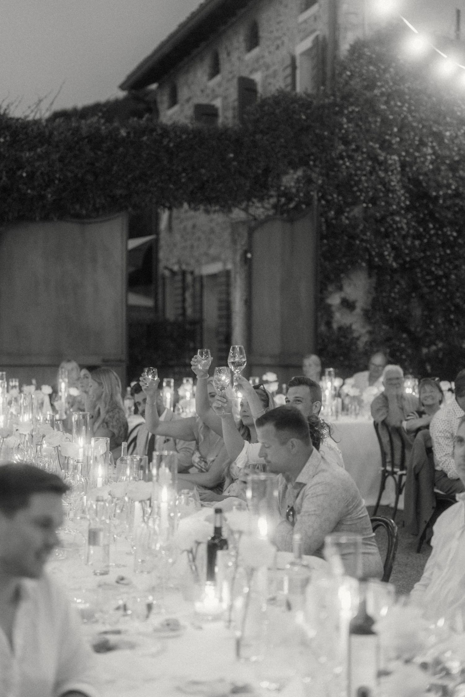 Black and white wedding reception dinner with guests raising glasses in candlelit toast at rustic venue.